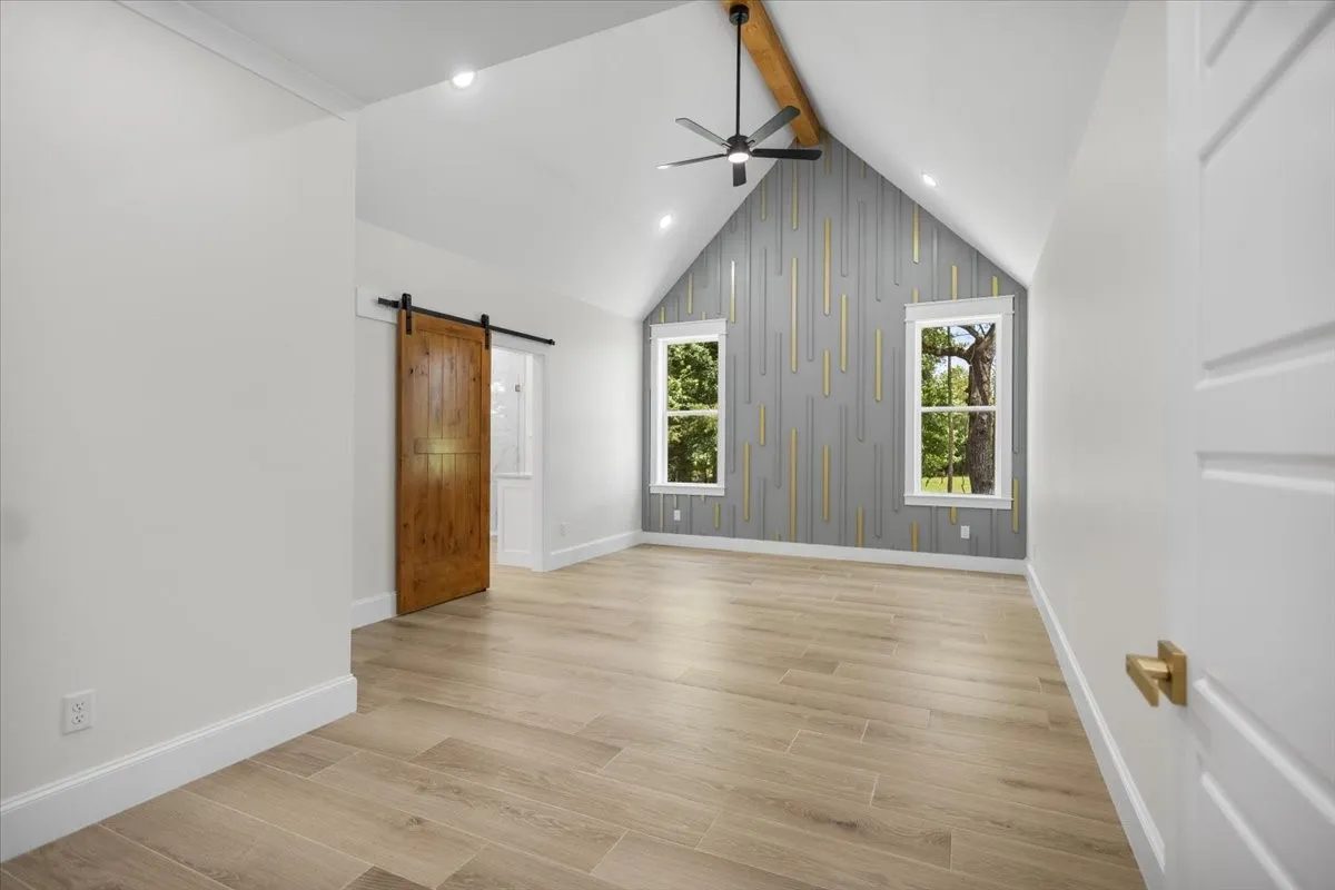 Vaulted ceiling with exposed wood beam, wood-finish flooring, two window openings, custom accent wall paneling, and barn door entry