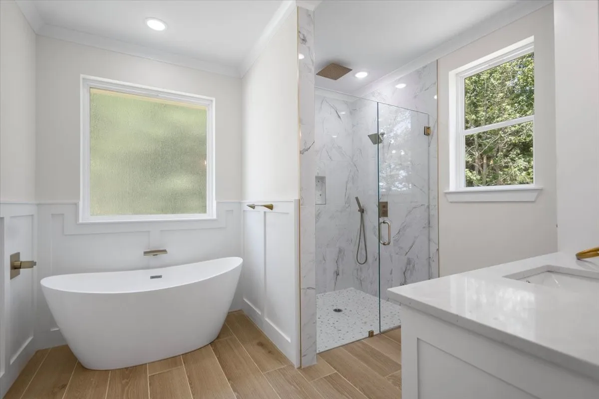 Freestanding soaking tub, walk-in shower with glass enclosure, wood-finish flooring, white vanity with integrated sink, and recessed lighting