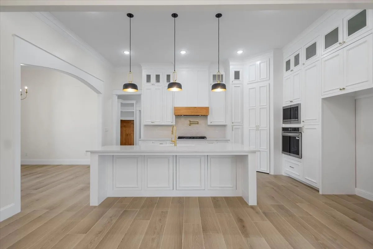 Expansive kitchen island with a light-toned countertop, integrated double wall ovens, abundant white cabinetry, and wood-finish flooring