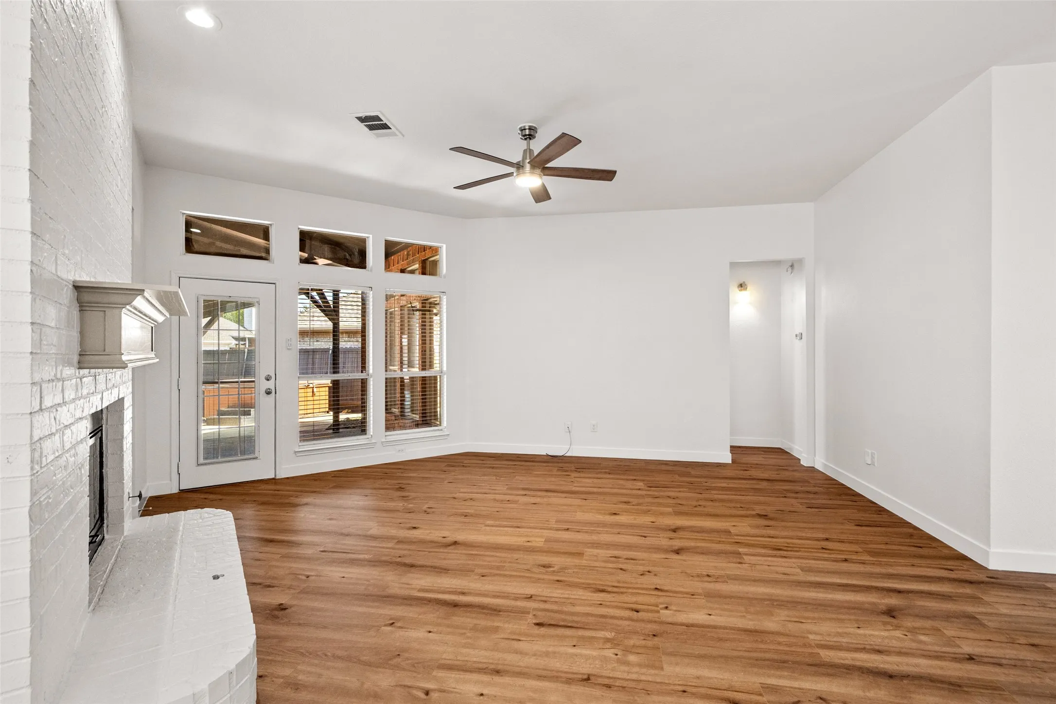 Wood-finish flooring throughout, white painted brick fireplace with a mantle, white walls, ceiling fan, and multiple windows