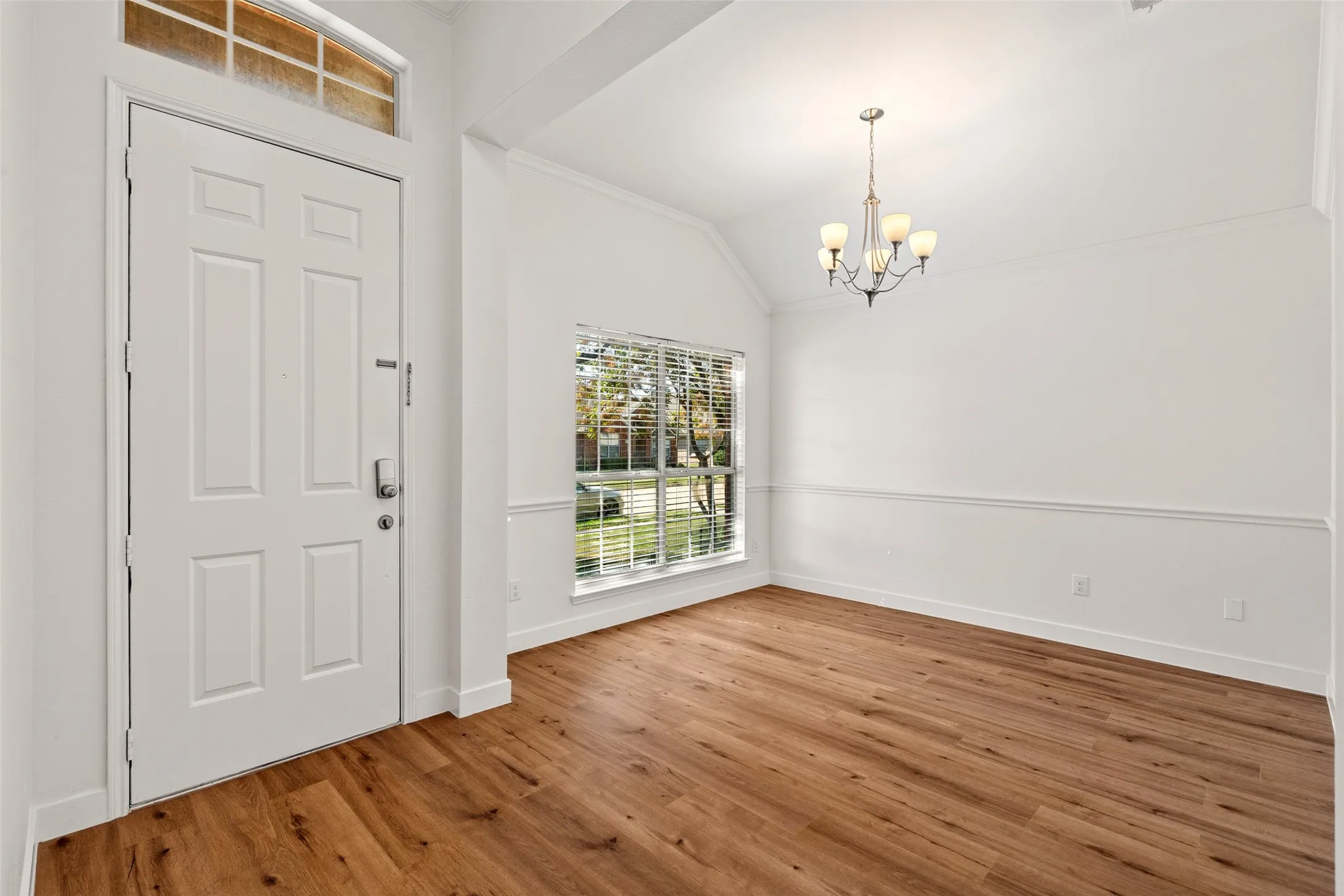Warm wood-finish flooring, white paneled door with transom window, window with horizontal blinds, decorative wall trim, and a classic chandelier
