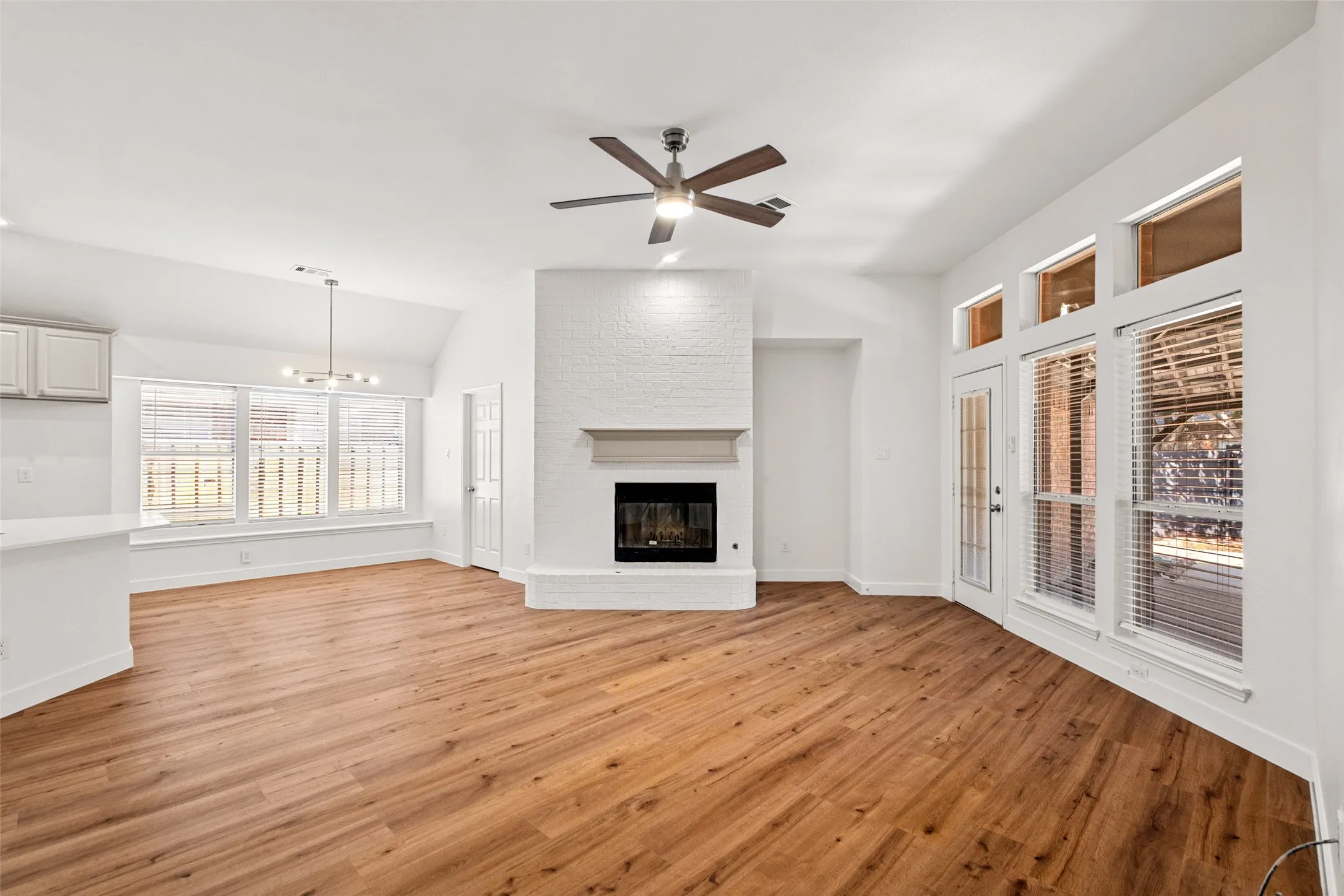 Spacious living area featuring wood-finish flooring, a white brick fireplace with a mantel, and an exposed beam ceiling fan