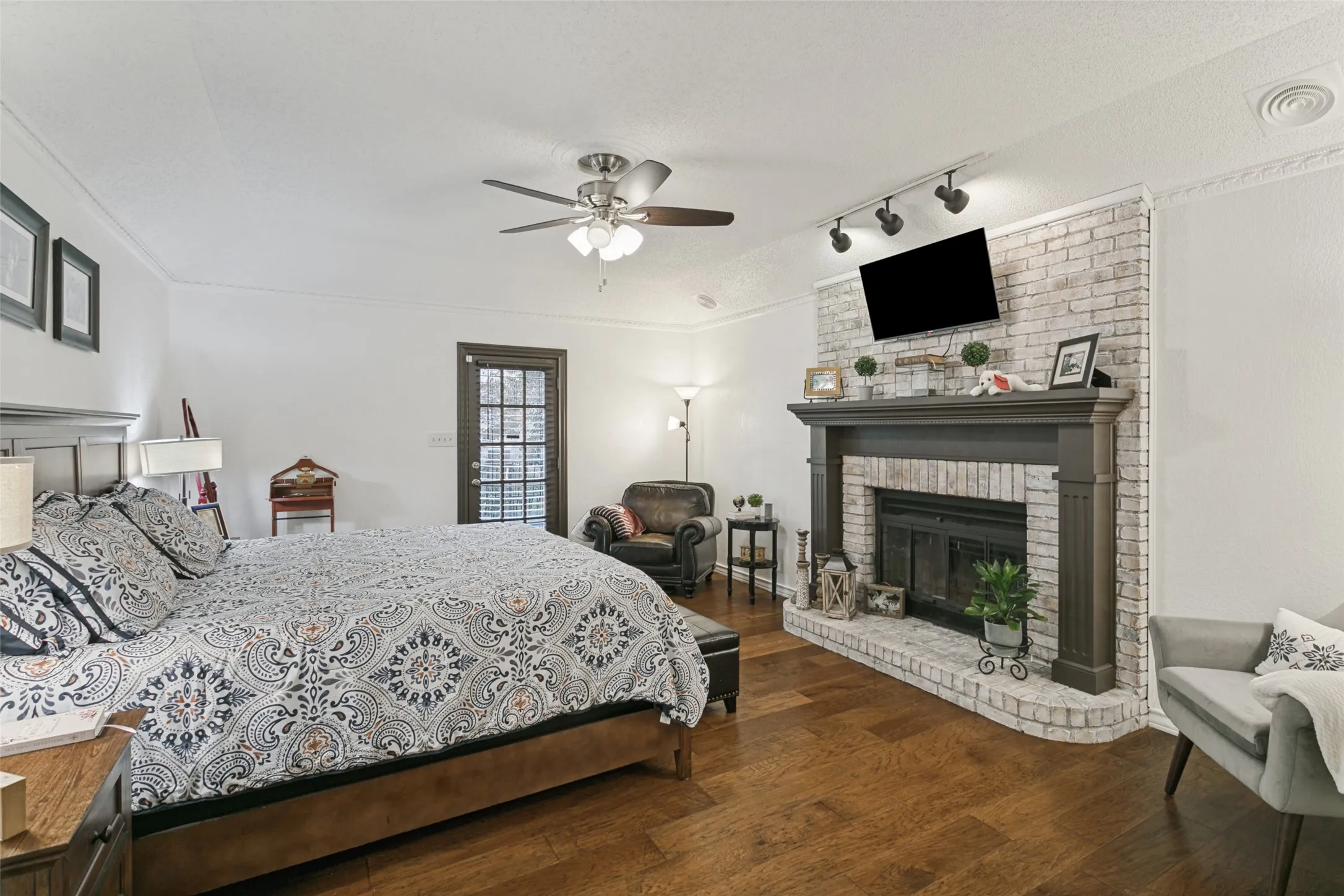 Primary bedroom with Rich wood-finish flooring, a white brick fireplace with a dark wood mantel, a ceiling fan, crown molding, and a paneled glass door
