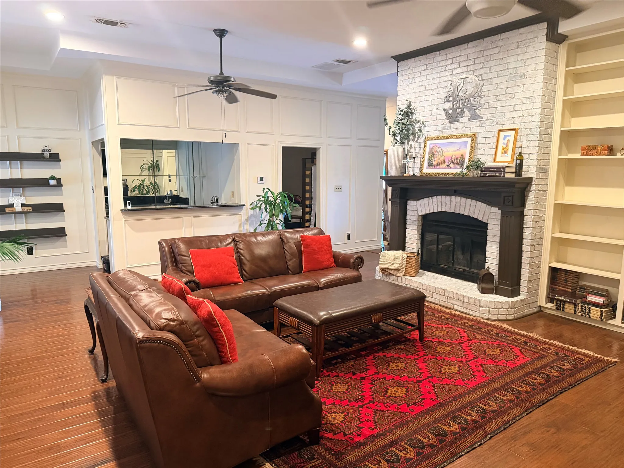 Wood-finish flooring, white brick fireplace with dark wood mantel, paneled walls, and a recessed ceiling with a ceiling fan