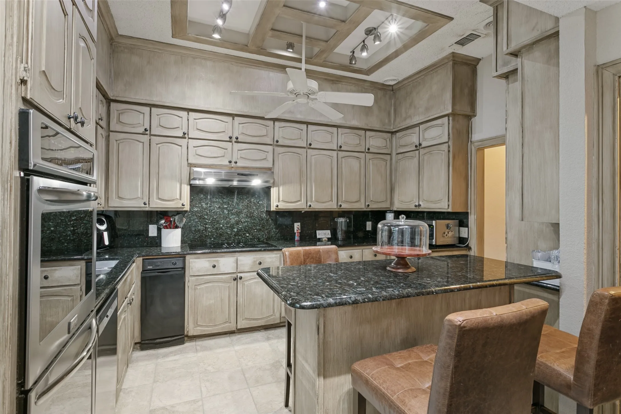 Kitchen featuring a coffered ceiling, stainless steel double wall oven, built-in dishwasher, dark stone countertops, and light-toned cabinetry