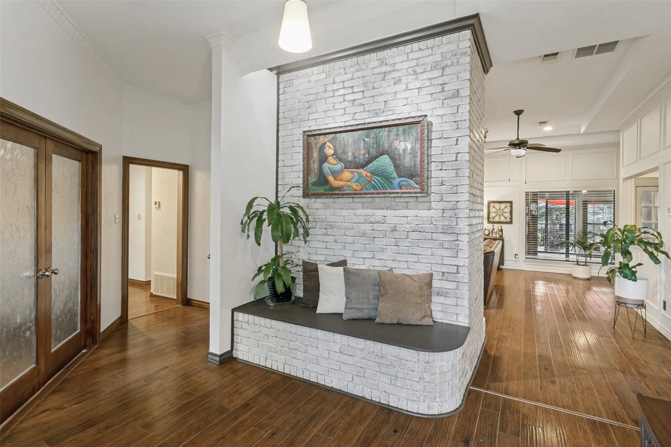 Wood-finish flooring throughout, white brick partition wall with built-in seating, French doors with frosted glass panels, crown molding, and a ceiling fan with integrated lighting