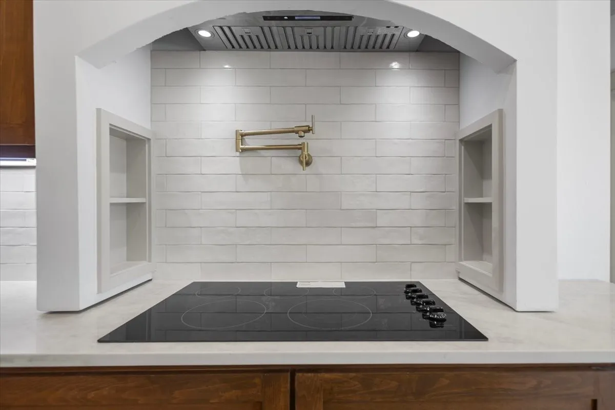 Cooktop with integrated controls, wall-mounted pot filler, tile backsplash, built-in recessed shelving, and under-cabinetry