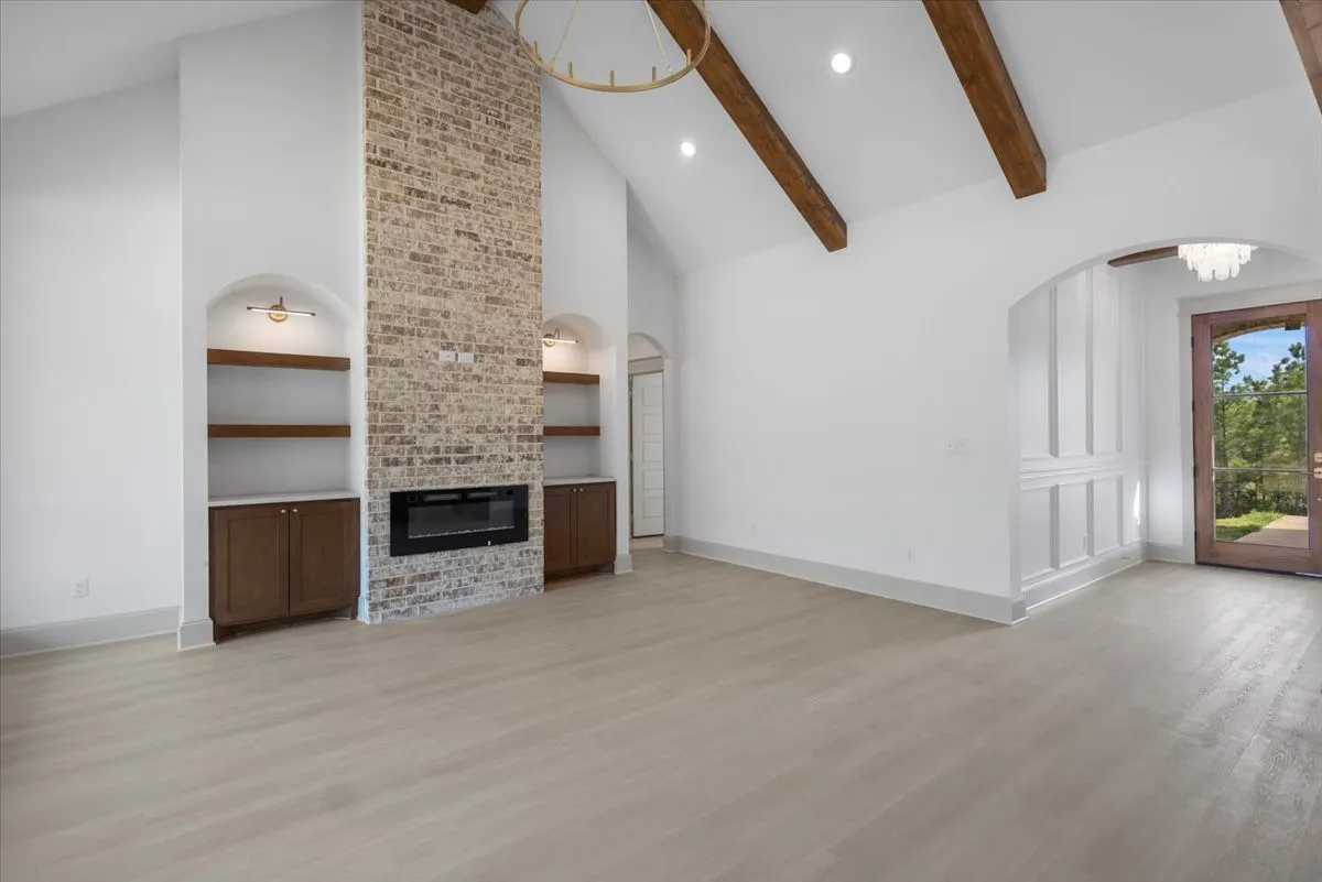 Vaulted ceiling with exposed wood beams, brick fireplace with an electric insert, built-in shelving with integrated cabinetry, wood-finish flooring, and an arched entry with wainscoting