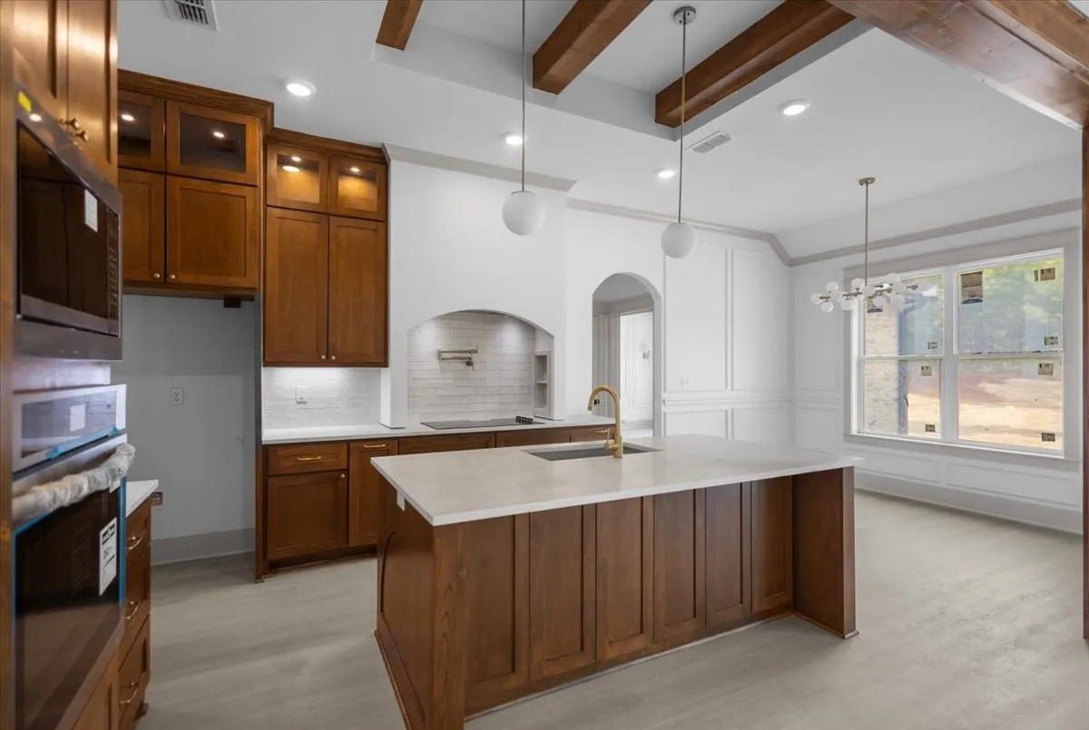 Hardwood cabinetry with integrated lighting, a large kitchen island with a light-toned countertop, a tile backsplash, and a decorative arched entryway