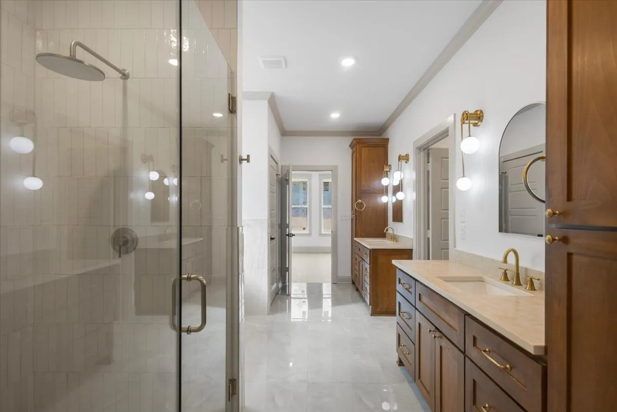 Spacious bathroom featuring a glass-enclosed shower with a rainfall showerhead, dual wood-finish vanities with integrated sinks and light-colored countertops, and gold-finish hardware throughout