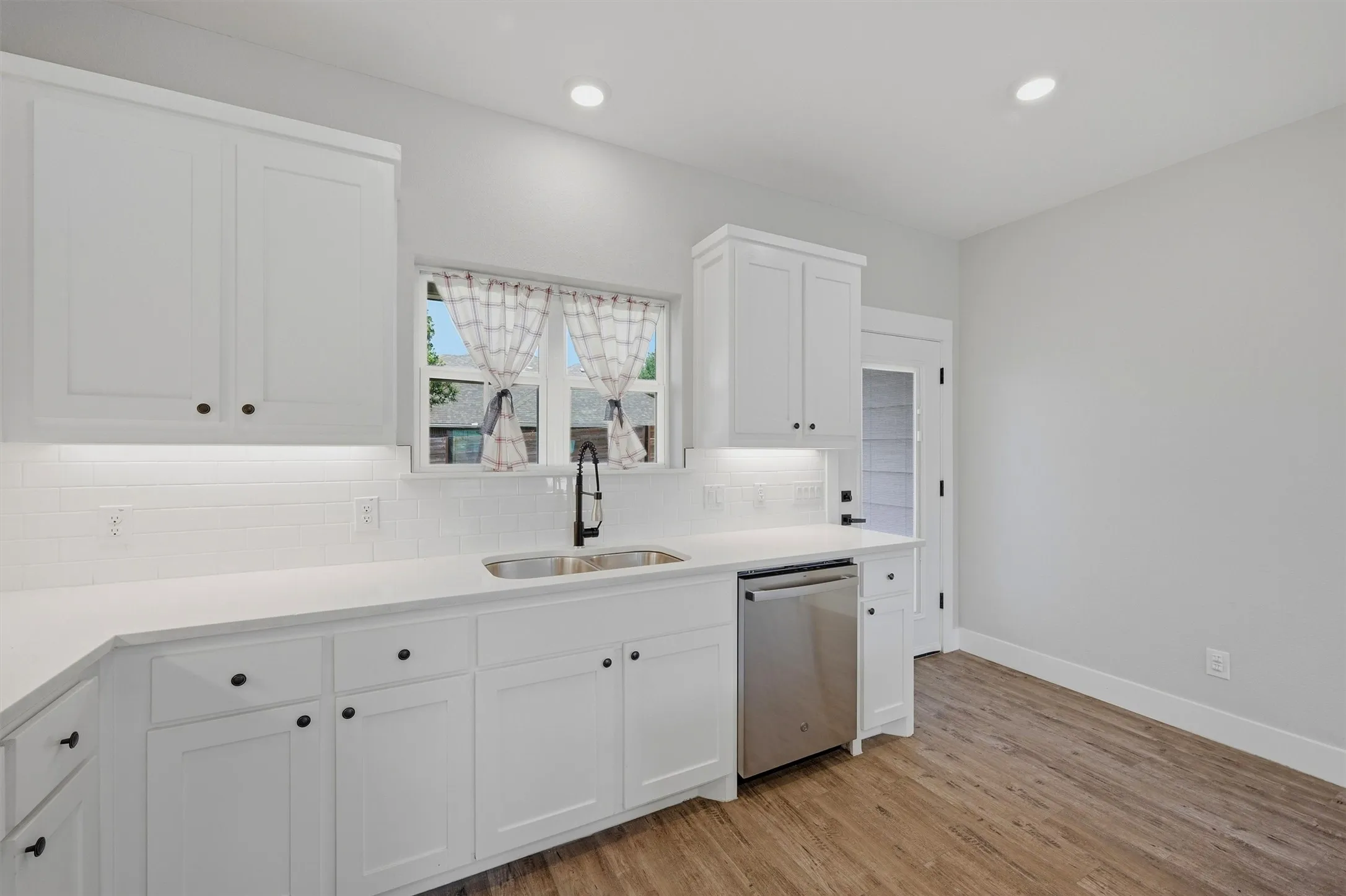 White cabinetry with black hardware, white subway tile backsplash, stainless steel sink with a black pull-down faucet, stainless steel dishwasher, and wood-finish flooring