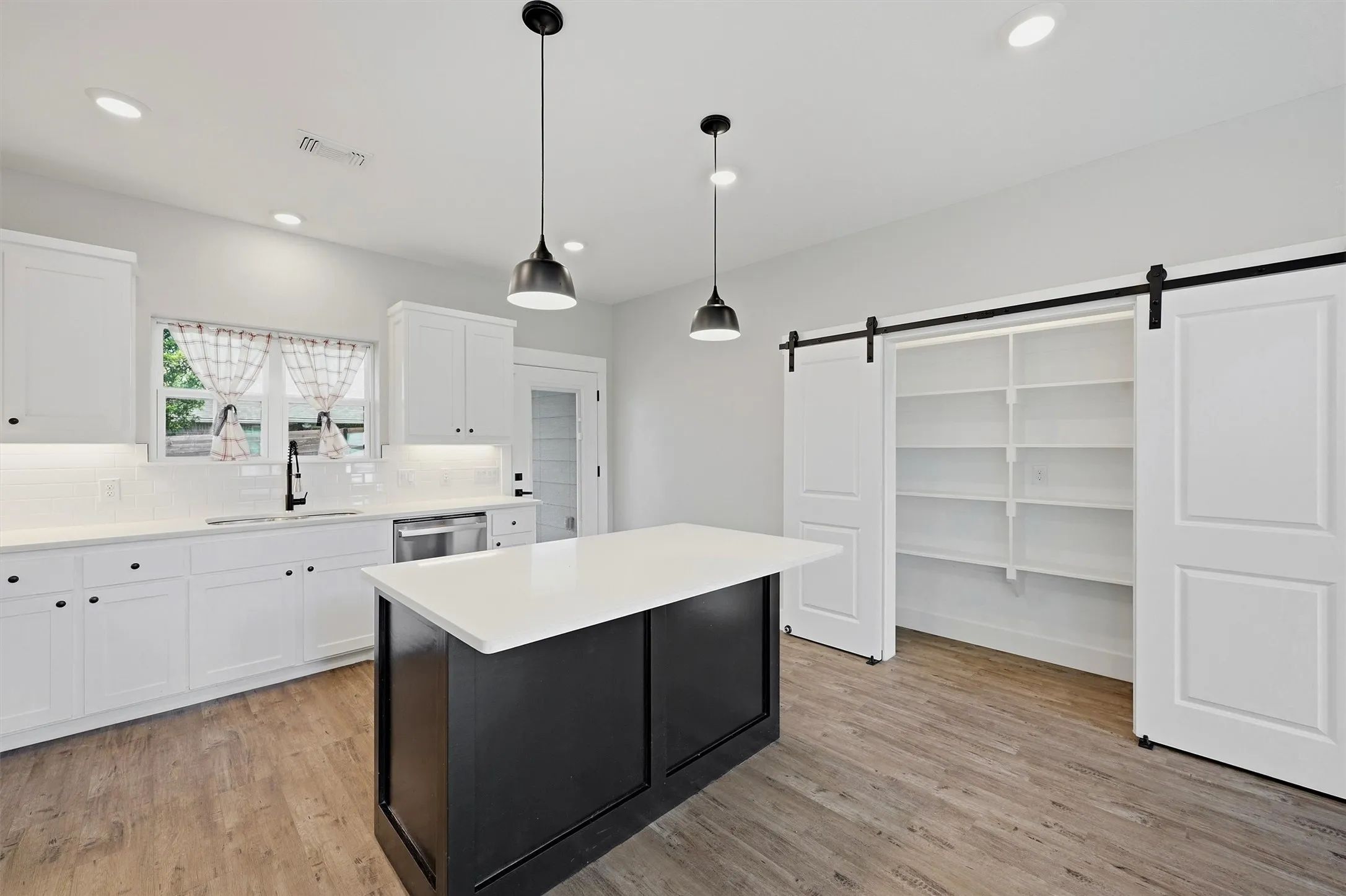 Kitchen featuring a central island with a light-colored countertop, white shaker-style cabinetry, wood-finish flooring, a stainless steel dishwasher, and a pantry with barn doors