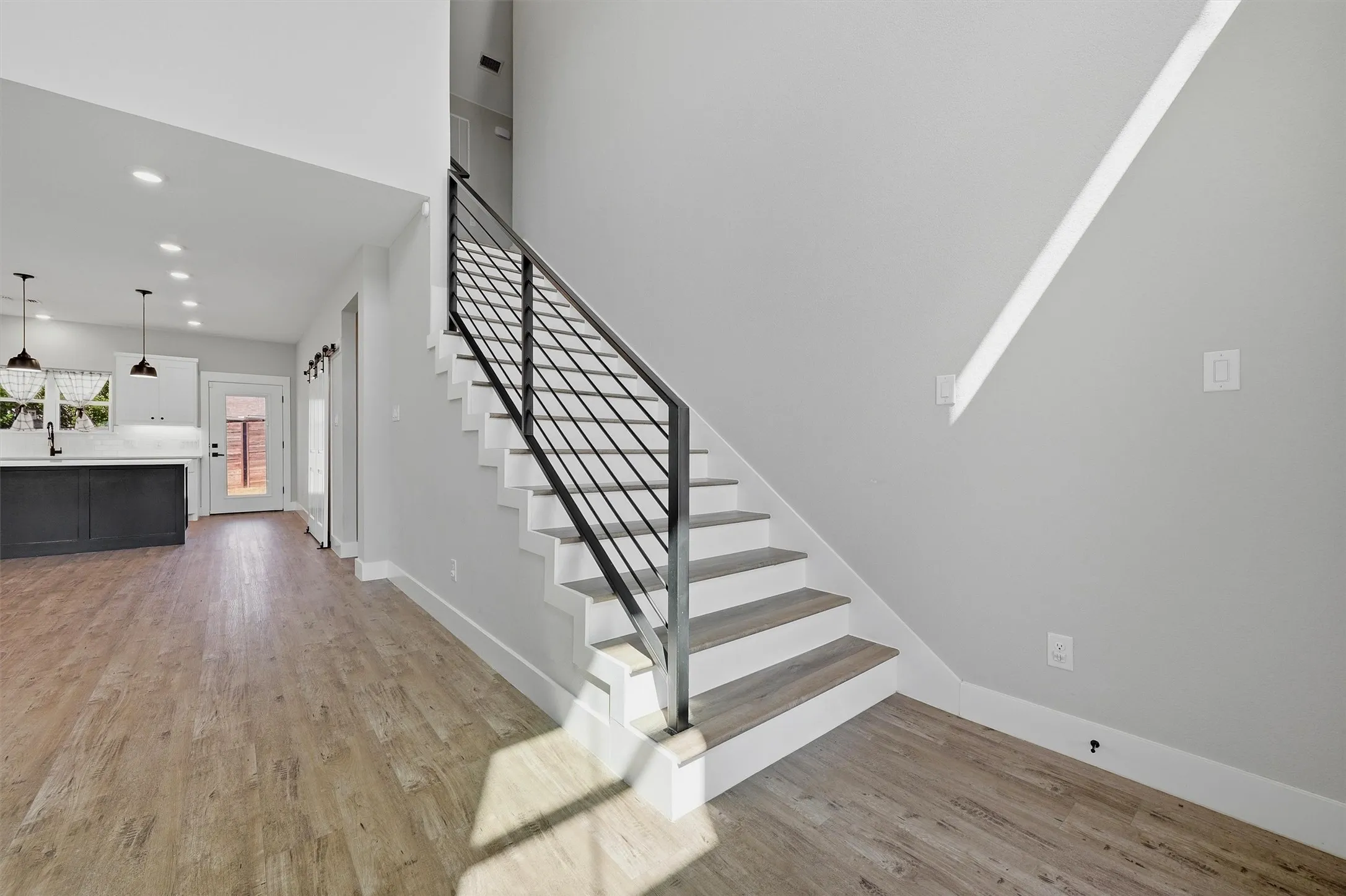 Modern staircase with black metal railing, wood-finish flooring, recessed lighting, and a white kitchen with a dark island