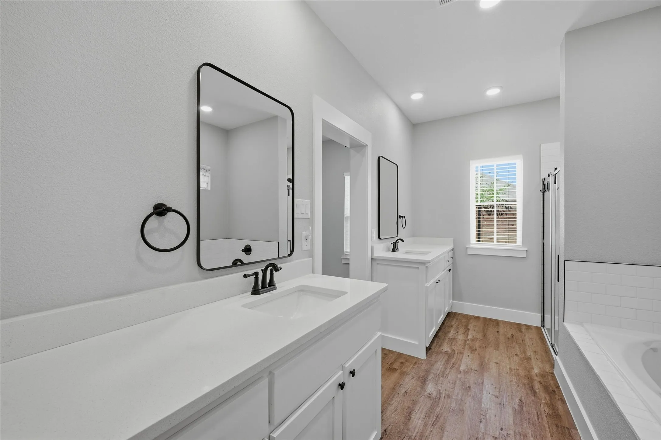Dual vanity with white cabinetry and countertops, matte black hardware, wood-finish flooring, and a separate tub and shower