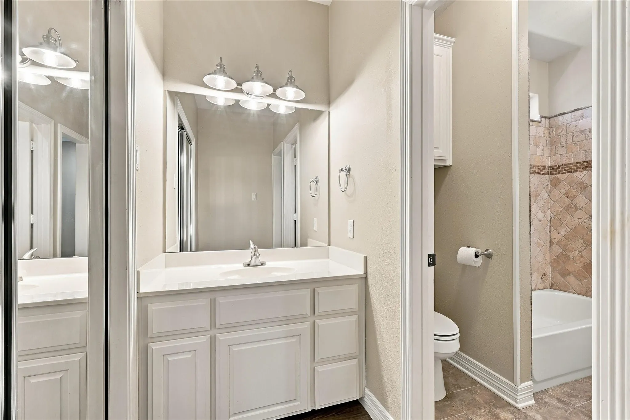 White vanity with integrated sink and chrome faucet, large vanity mirror with a four-light fixture, tile flooring, and a bath with patterned tile surround