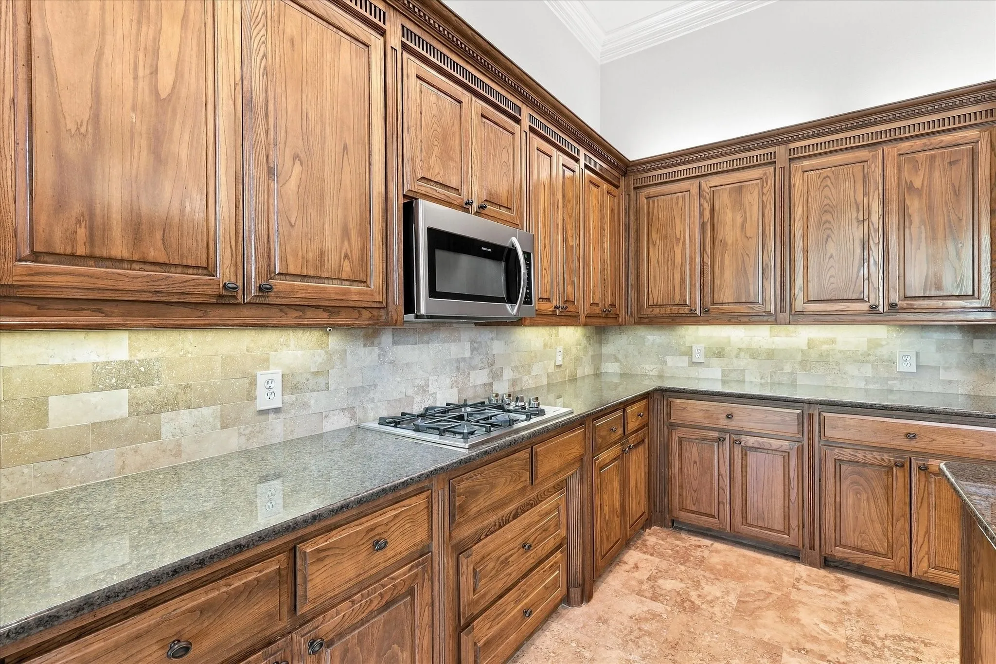 Extensive wood cabinetry, stainless steel microwave, gas cooktop, stone tile backsplash, and light-toned flooring