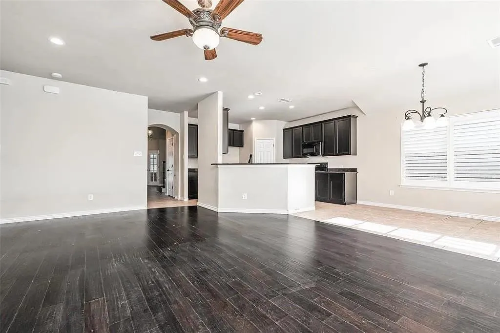 Dark wood-finish flooring, recessed ceiling lighting, ceiling fan with light fixture, kitchen with dark cabinetry and countertops, and window with plantation shutters