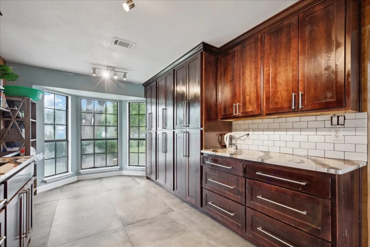 Bay window with multiple panes, extensive cabinetry with dark wood finish, light-toned tile flooring, subway tile backsplash, and light-toned countertop