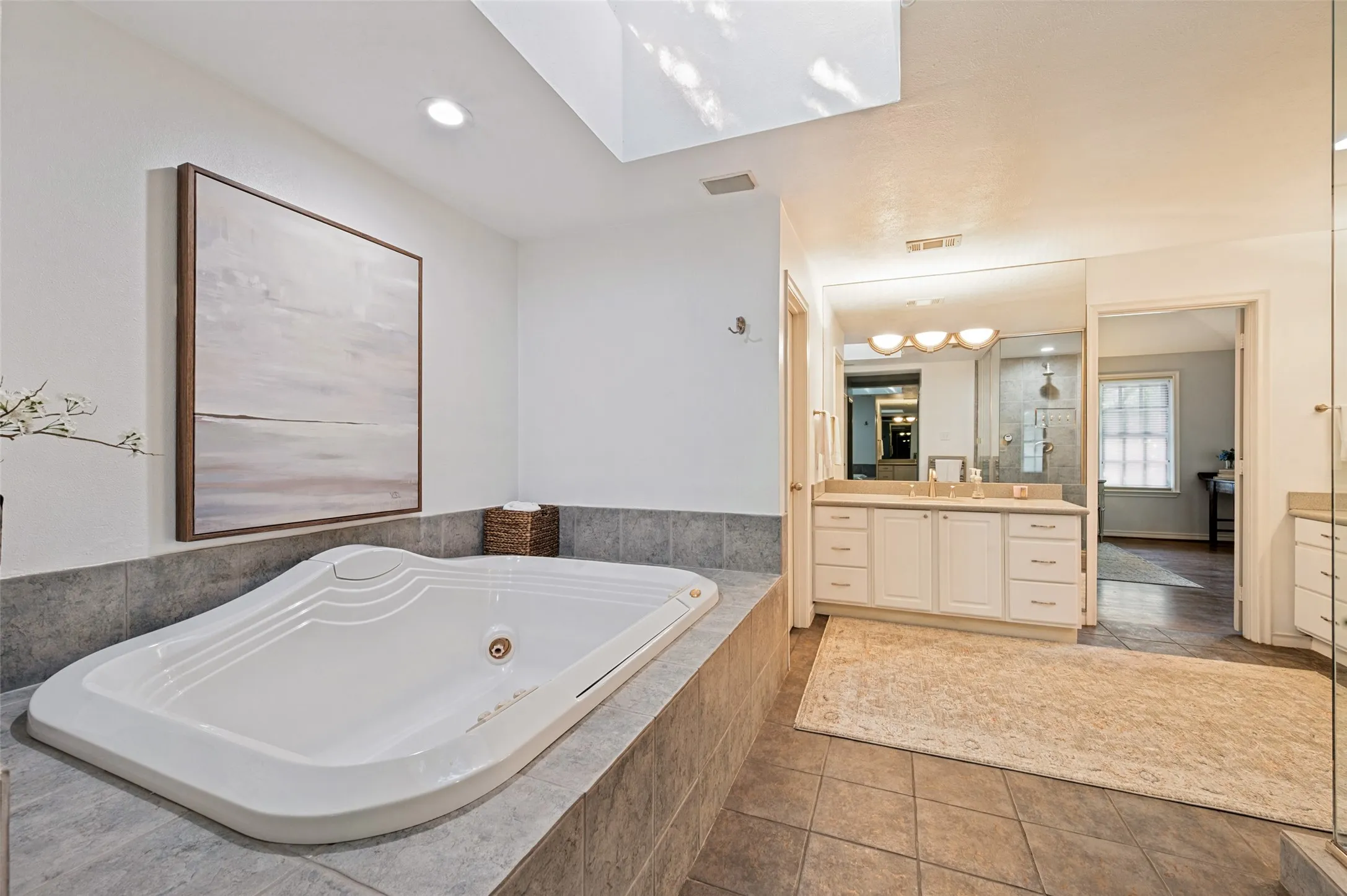Soaking tub with tiled surround, dual vanity with undermount sinks, and a large mirror with integrated lighting
