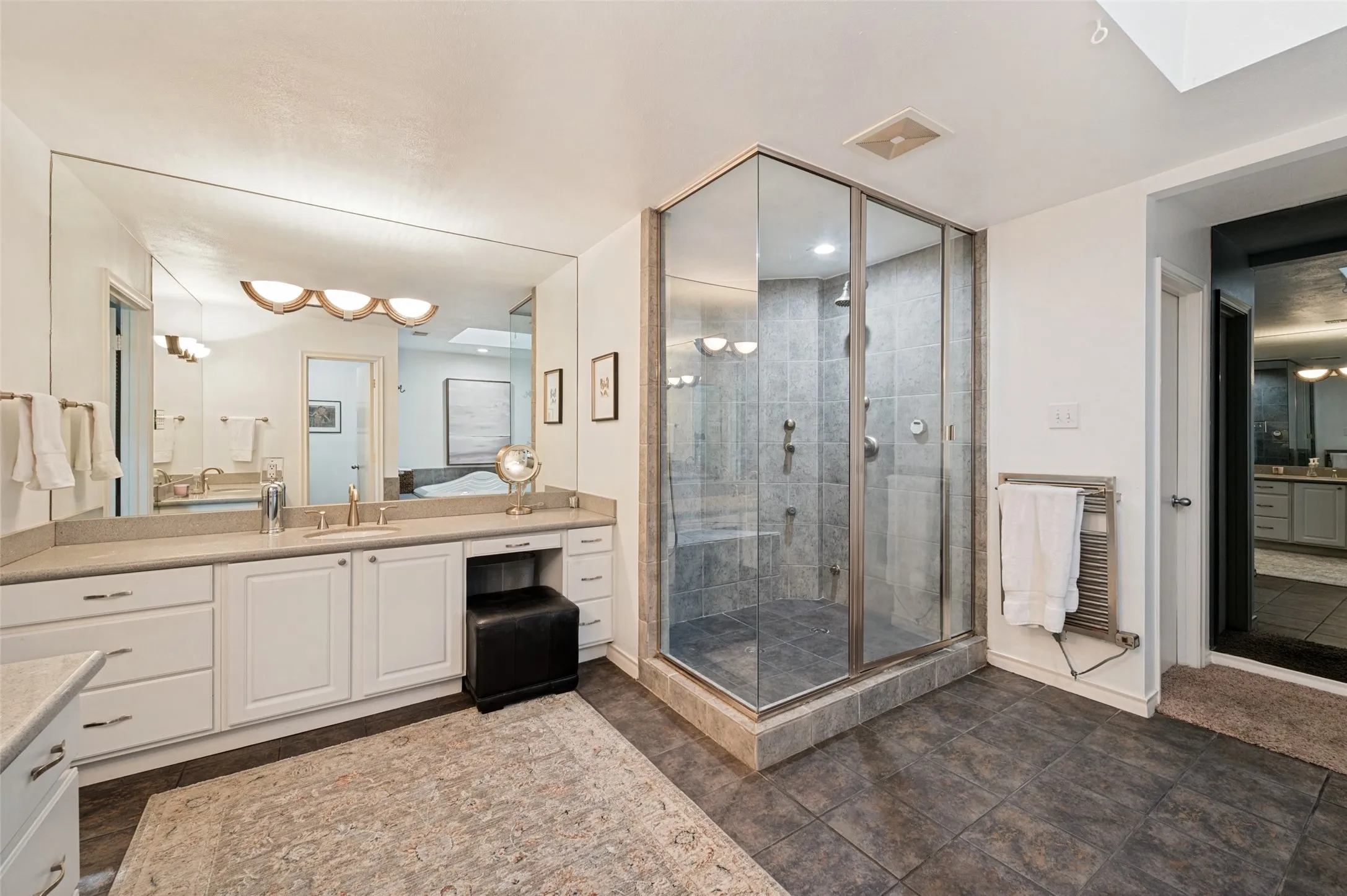 Glass-enclosed shower with a built-in bench, dual vanity with white cabinetry, large wall-mounted mirror, and tile flooring