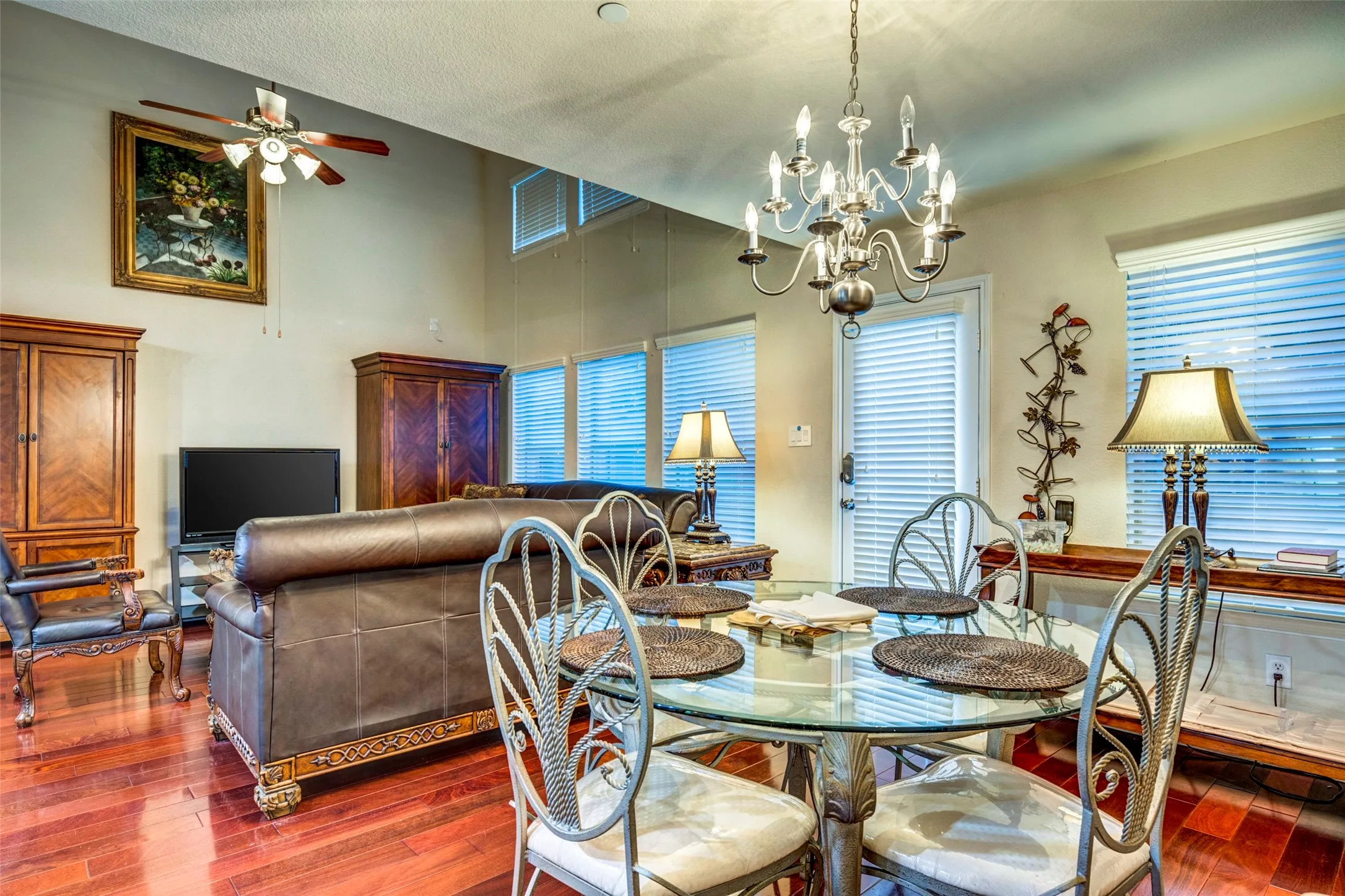 Open-concept living area with high ceilings, wood-finish flooring, a contemporary chandelier, and multiple windows with horizontal blinds
