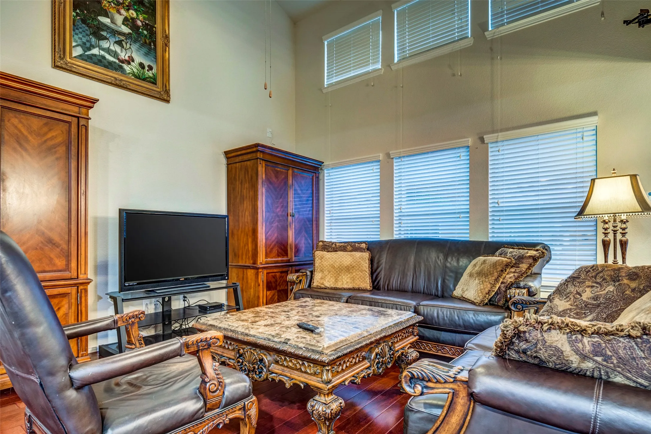 Sophisticated living room showcasing soaring ceilings with transom windows, ornamental wood cabinetry with glass-front doors, elegant upholstered seating, decorative wood-finish flooring, and table lamps with brass accents