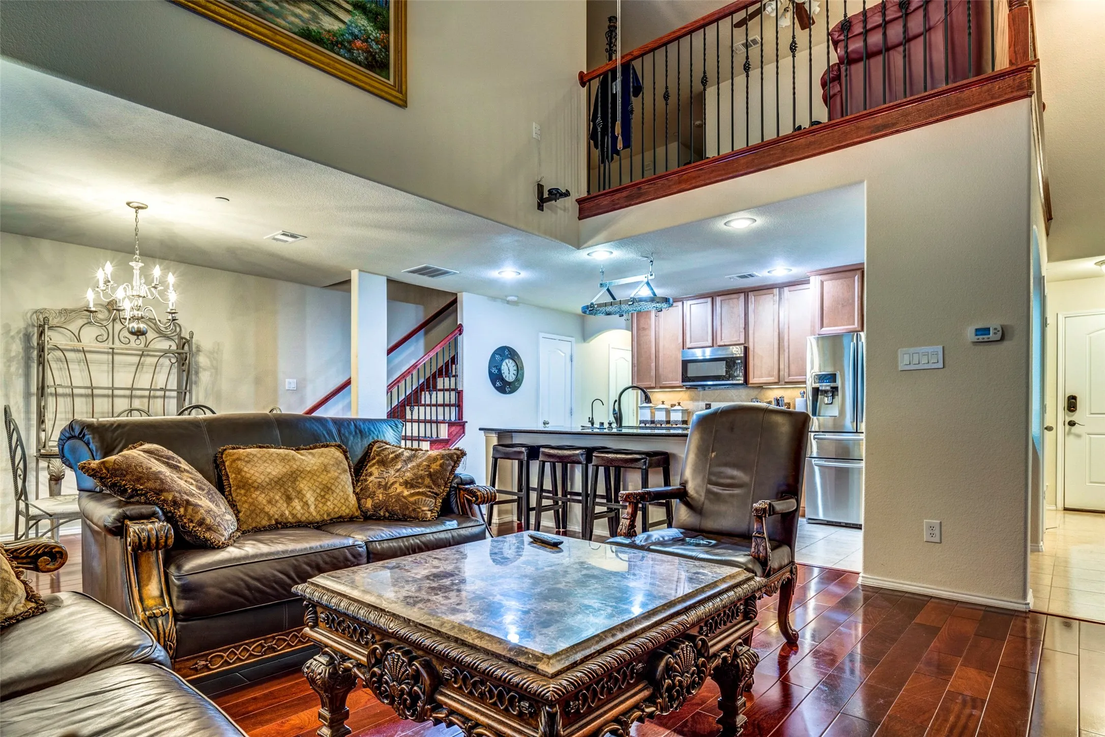 Two-story living area with wood-finish flooring, an upper-level balcony, and a visible kitchen featuring stainless steel appliances and wood cabinetry