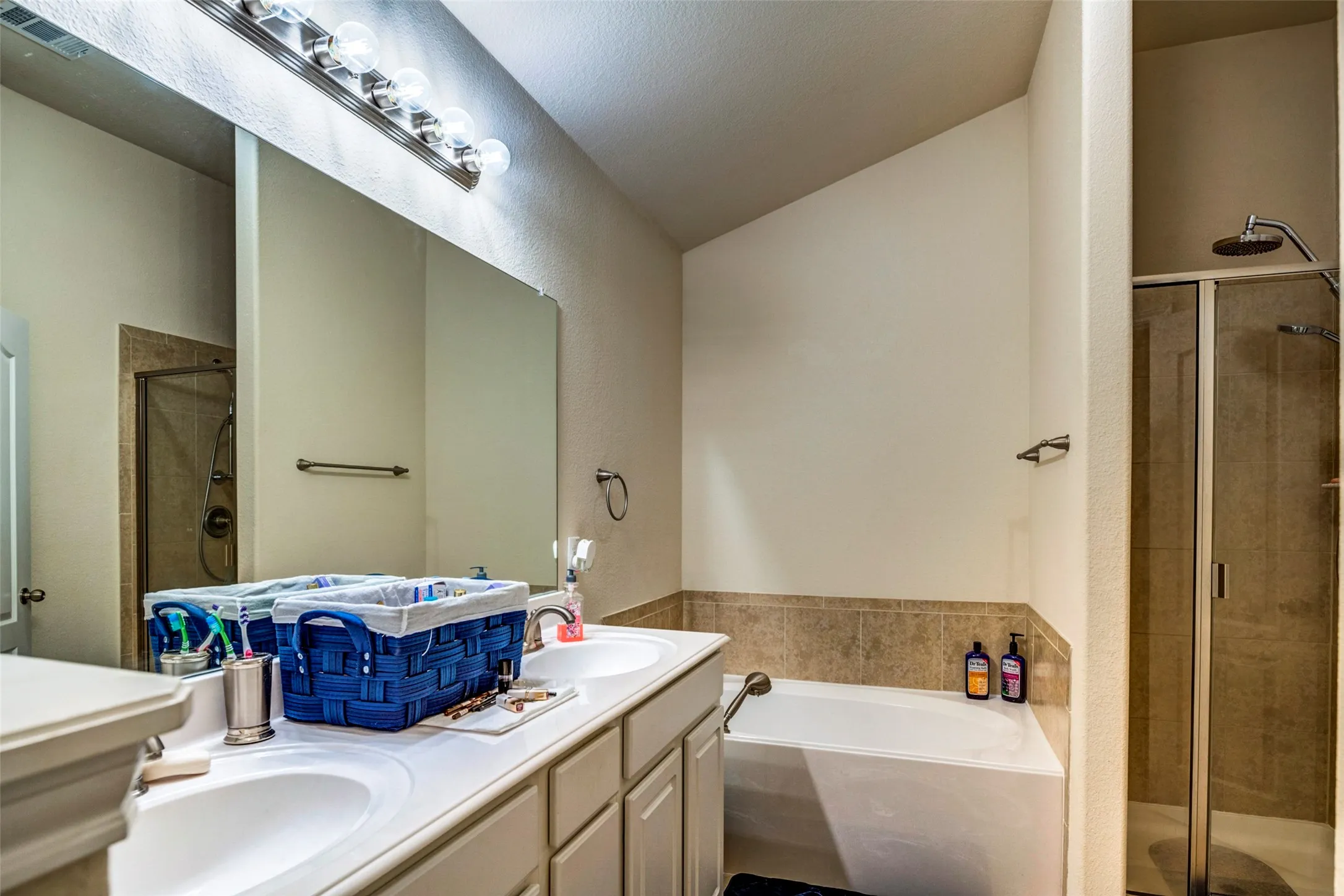 Master bathroom featuring dual vanity with wood-finish cabinetry and white countertop, large framed mirror with integrated lighting, soaking tub with tile surround, and separate walk-in shower with glass enclosure and tile detailing