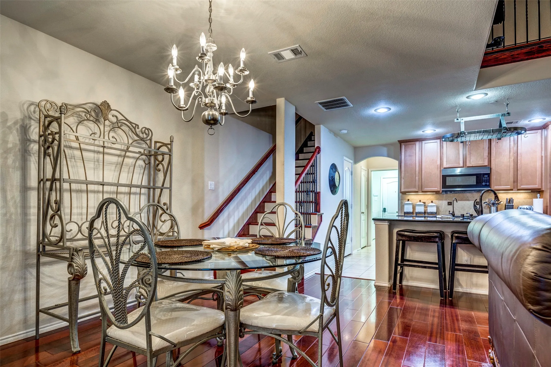 Open-concept dining and kitchen area featuring a round glass-top table with ornamental metal base and chairs, wood-finish flooring, pendant lighting, stainless steel appliances, wood cabinetry, and a curved archway entry