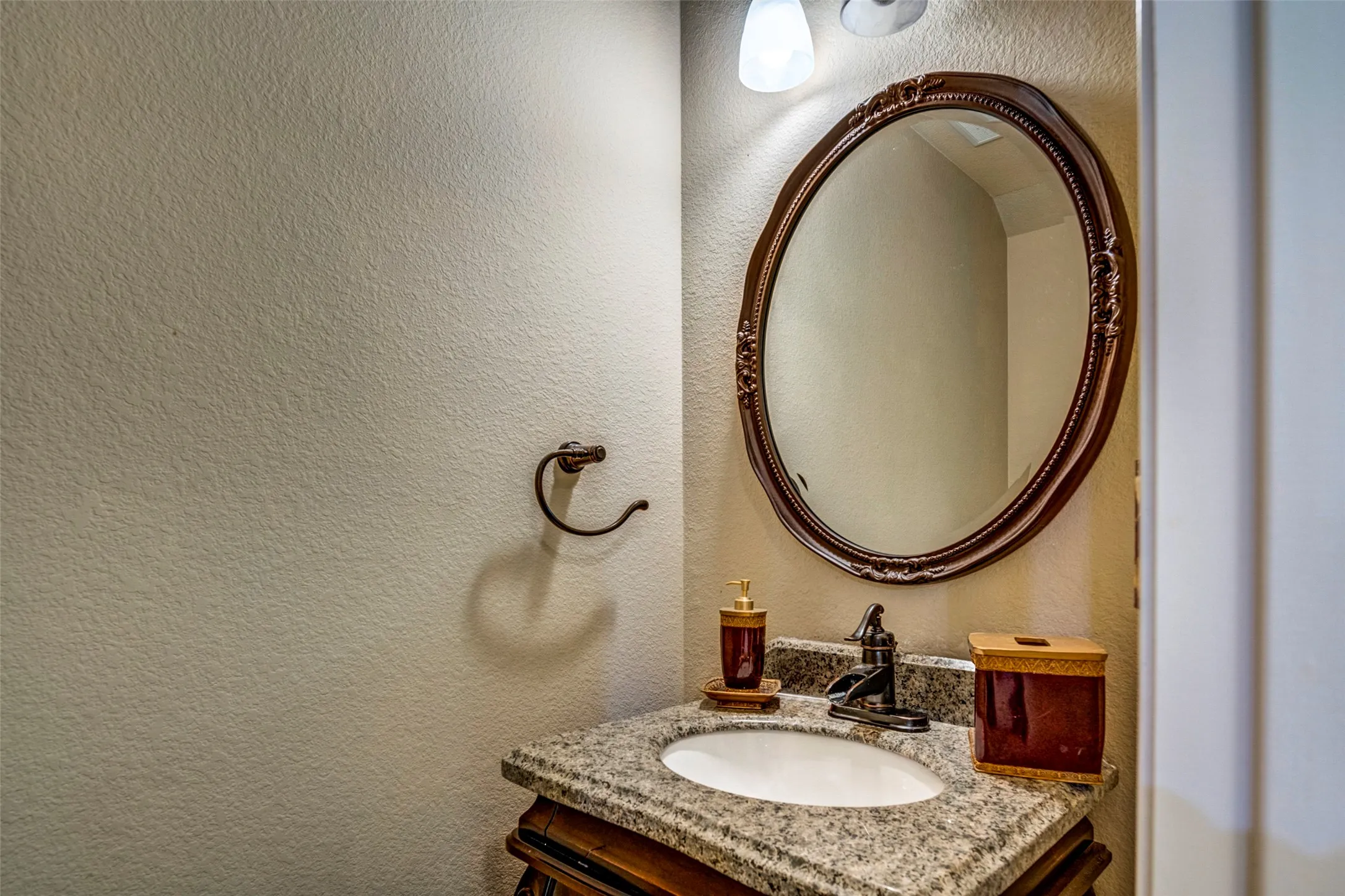 Oval mirror with an ornate frame, granite countertop, undermount sink, and wall-mounted vanity lighting