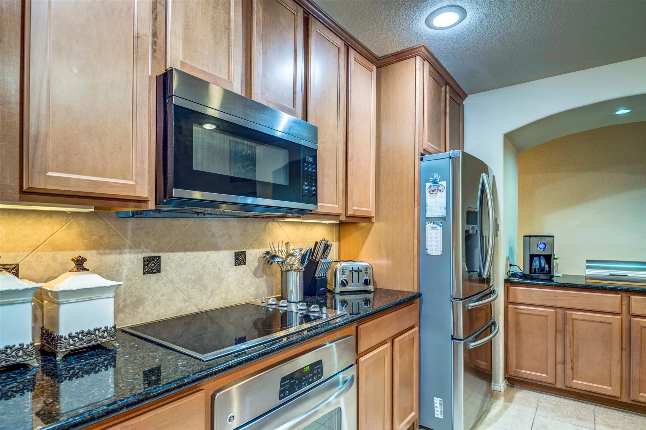 Updated kitchen featuring warm wood cabinetry, black granite countertops, stainless steel appliances including a microwave oven and French-door refrigerator, and a decorative tile backsplash with recessed lighting throughout