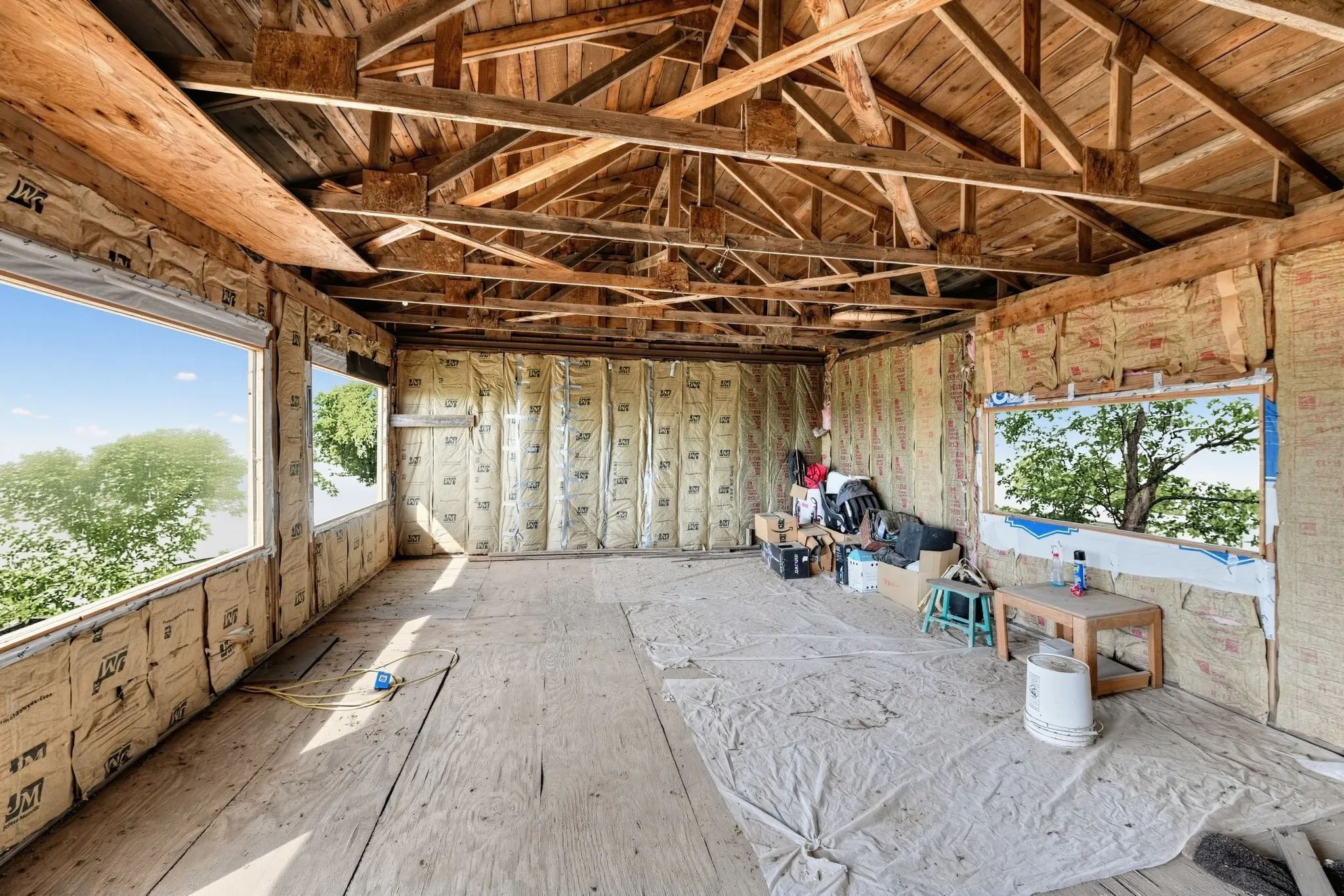Exposed wood beam ceiling structure, visible wall insulation, plywood subflooring, and multiple window openings