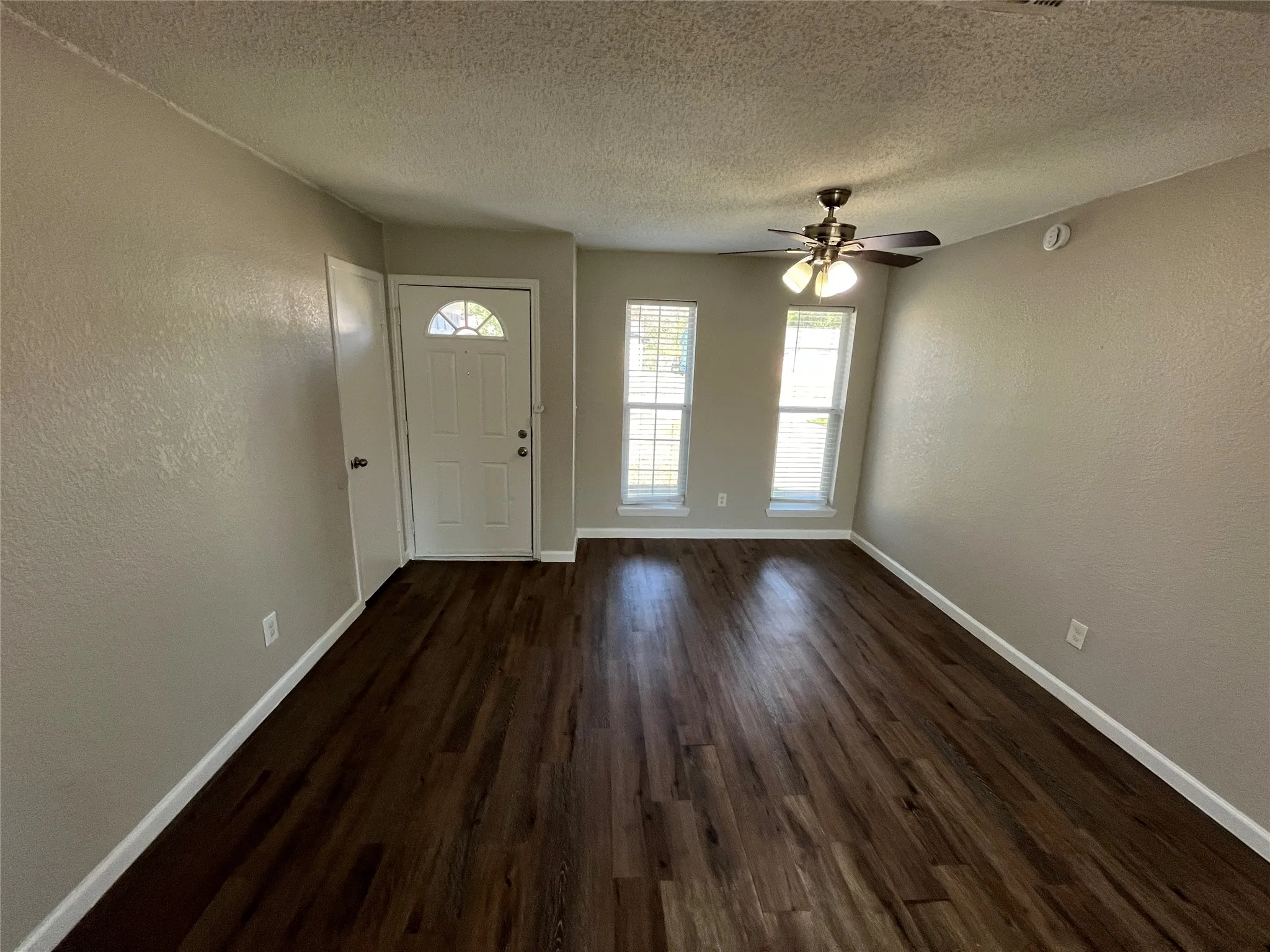 Spacious room with dark wood-finish flooring, light neutral-toned walls, multiple windows, and a ceiling fan with integrated lighting
