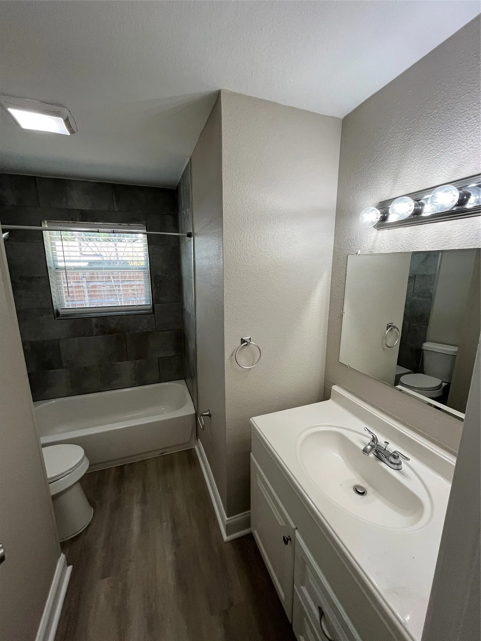 Bathroom featuring wood-finish flooring, a white vanity with an integrated sink, a built-in bathtub with dark tile surround, and a chrome light fixture