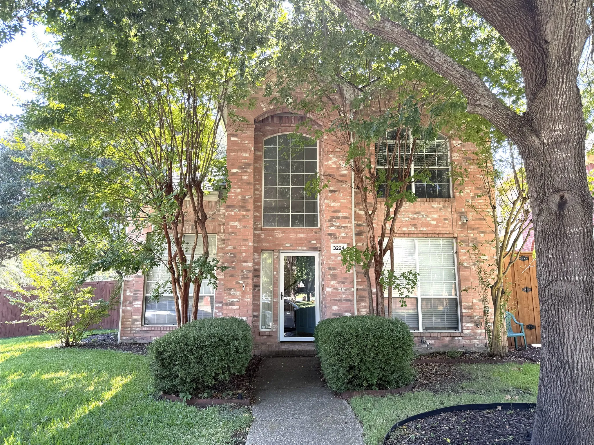 Brick exterior featuring an arched window, large picture windows, mature trees, manicured lawn, and a concrete pathway