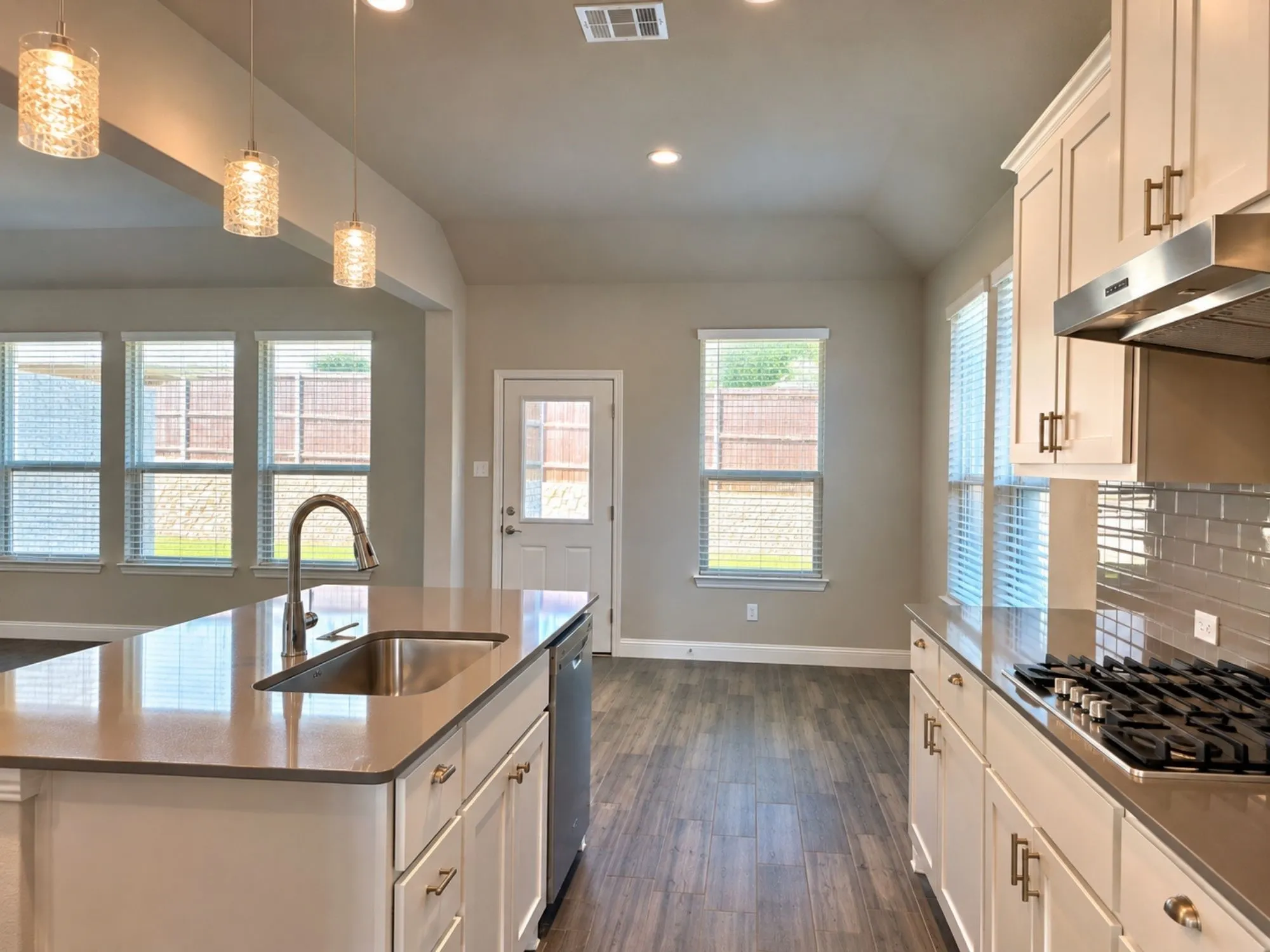 Galley-style kitchen featuring white cabinetry with brushed nickel hardware, stainless steel gas cooktop and dishwasher, subway tile backsplash, wood-finish flooring throughout, and pendant lighting above the center island with an undermount sink and pull-down faucet
