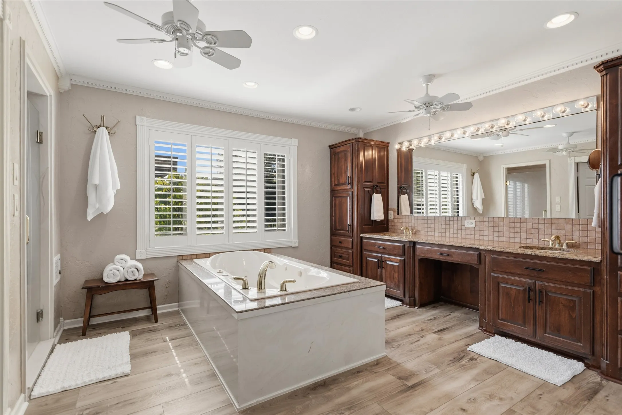 Soaking tub with surrounding deck, dual vanity with extensive counter space, warm wood-finish cabinetry, wood-finish flooring, and crown molding