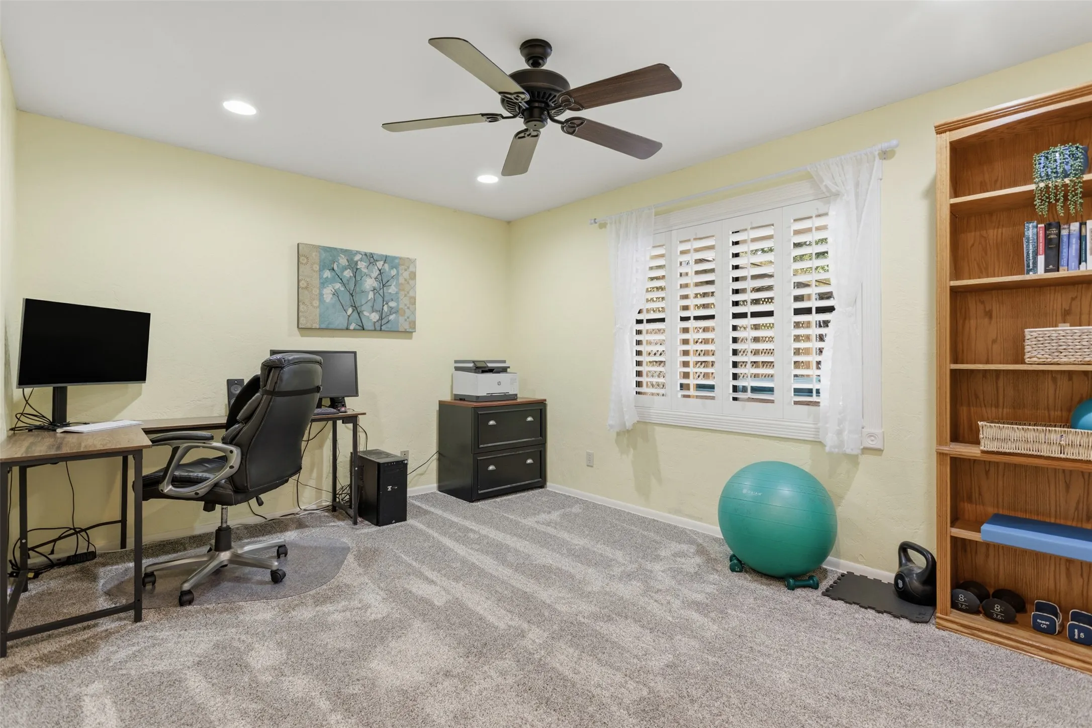 Versatile home office featuring a dedicated work desk, black five-blade ceiling fan with integrated lighting, soft yellow walls, and abundant natural light from shuttered windows