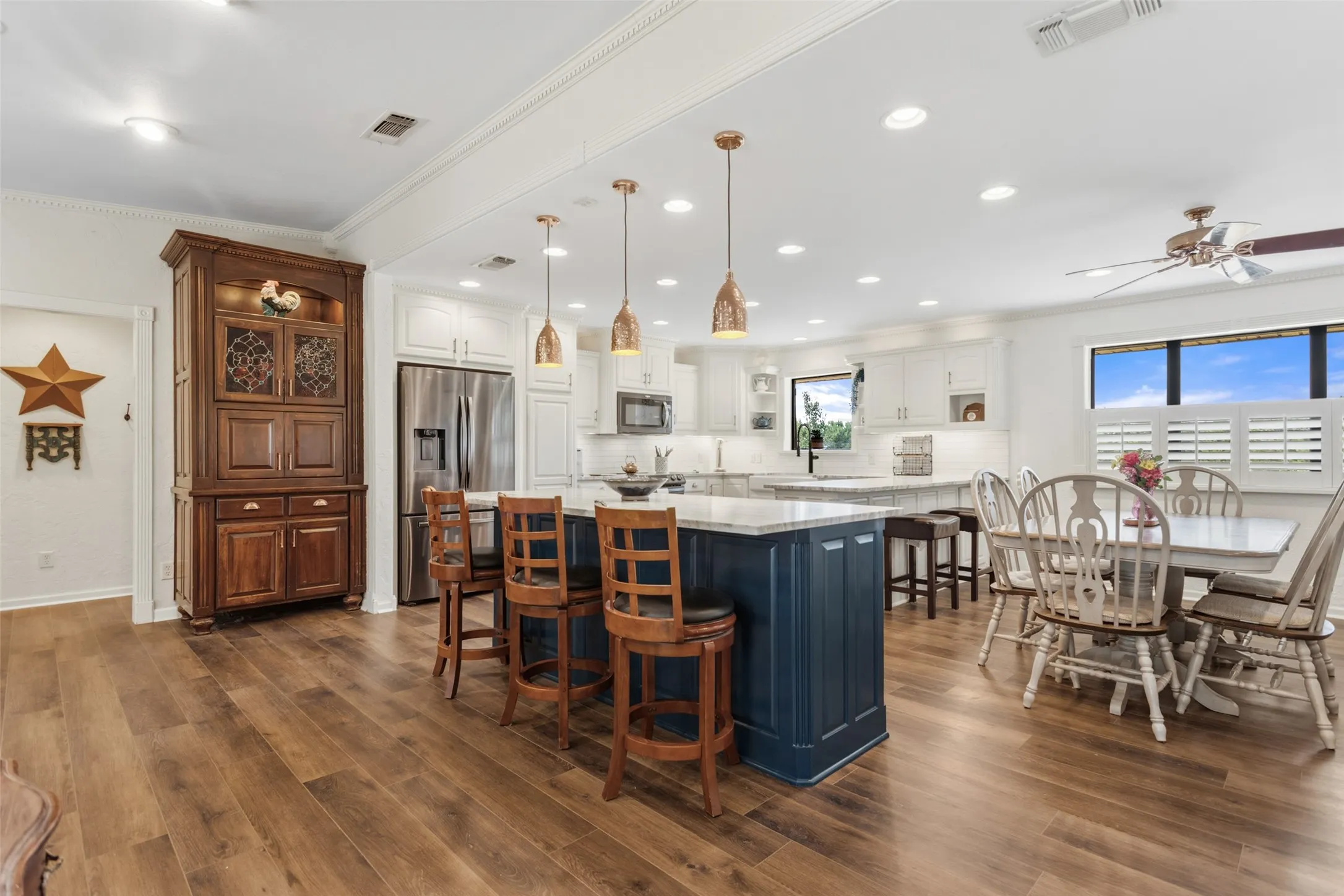 Open-concept kitchen and dining area featuring custom cabinetry with a navy island, stainless steel appliances, pendant lighting, wood-finish flooring throughout, and expansive windows with natural light