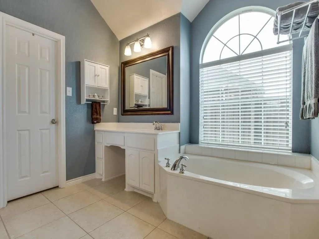 Vaulted ceiling, arched window with grilles, built-in bathtub with deck, white vanity with integrated sink, and large format tile flooring
