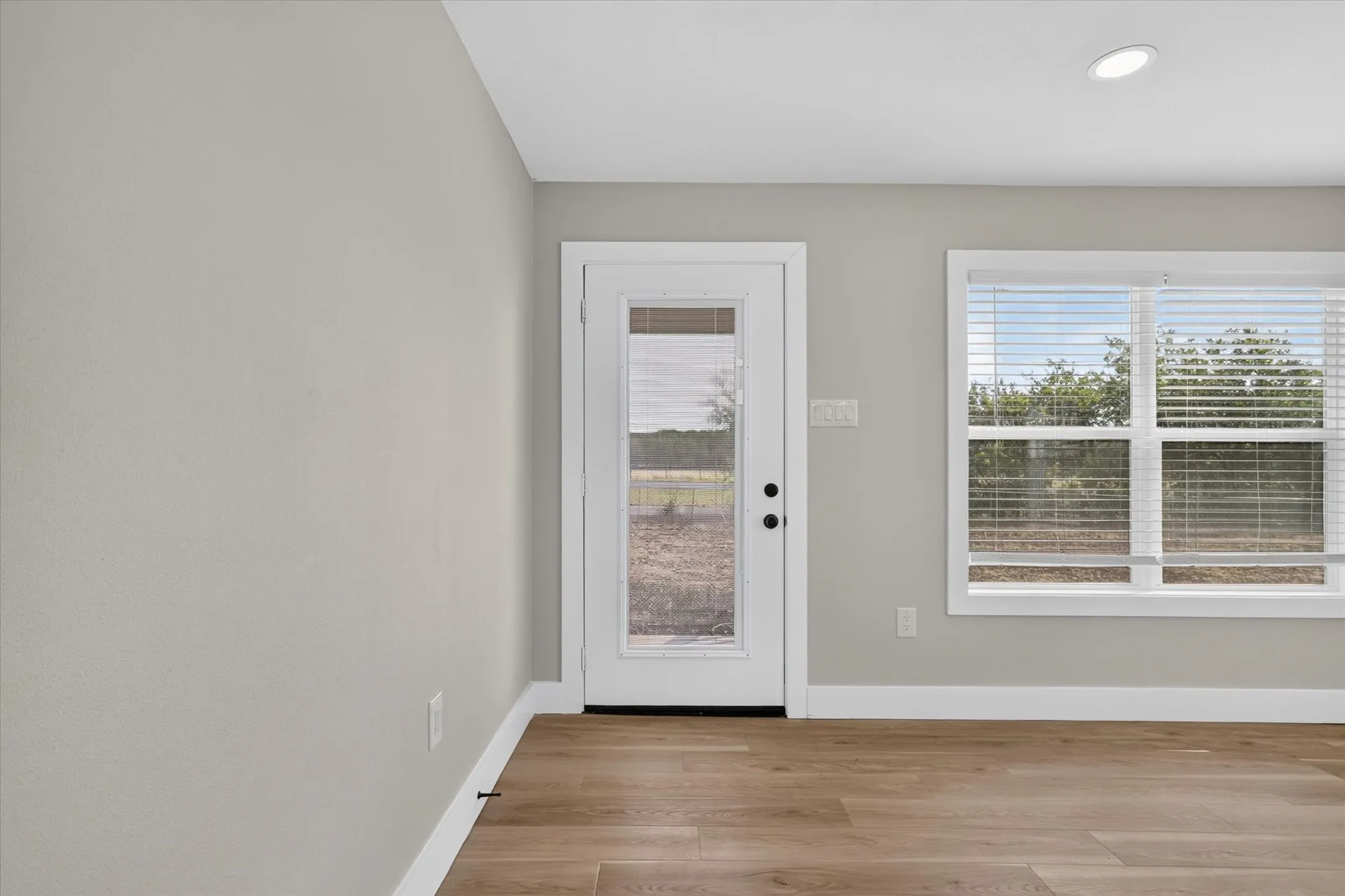 Bright and airy room showcasing gleaming wood-finish flooring, white trim detailing, and abundant natural light from a full-glass entry door and large dual-pane windows with integrated blinds