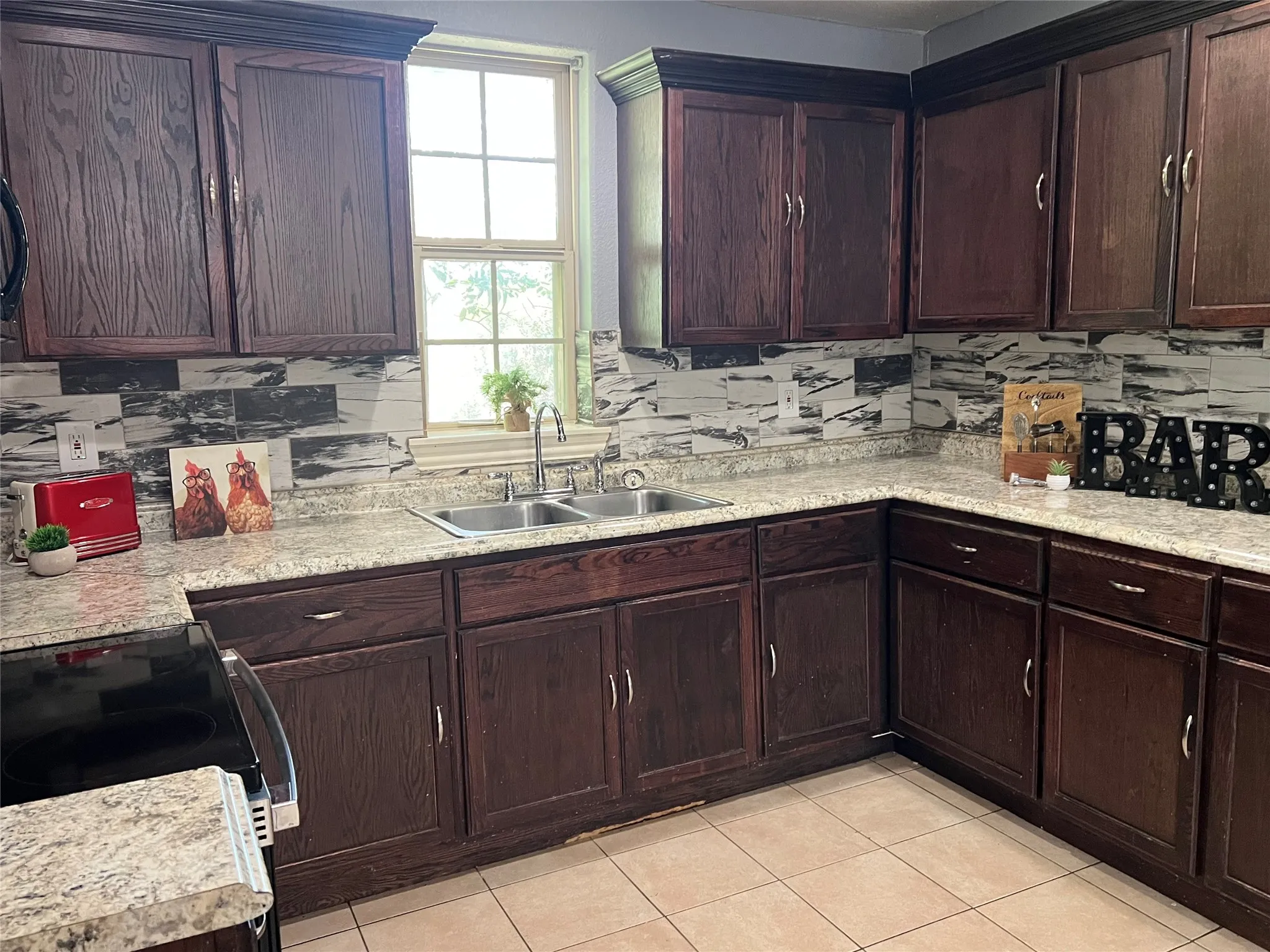 Dark wood-finish cabinetry with crown molding, light-toned countertops, tile backsplash, and a double basin sink with a gooseneck faucet