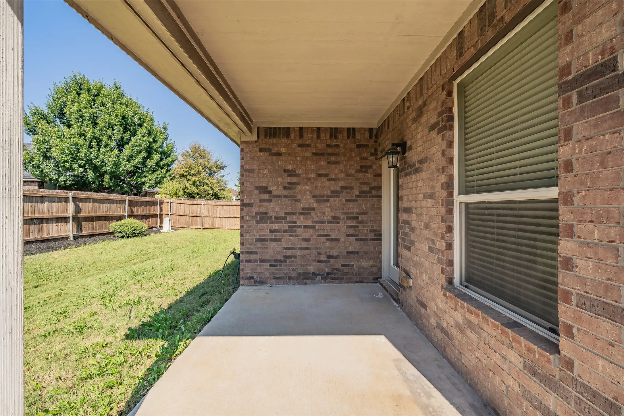 Covered concrete patio with a brick exterior, outdoor wall sconce, and a fenced backyard