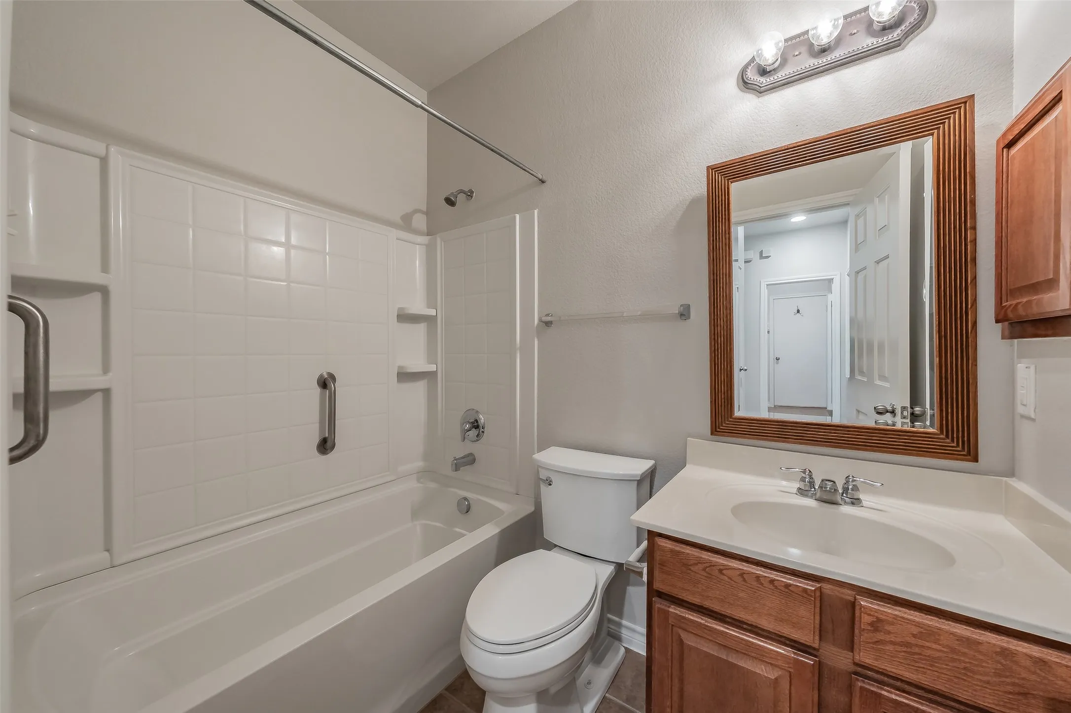 Built-in bathtub with white tile surround, wood-finish vanity with countertop, framed mirror, and chrome fixtures