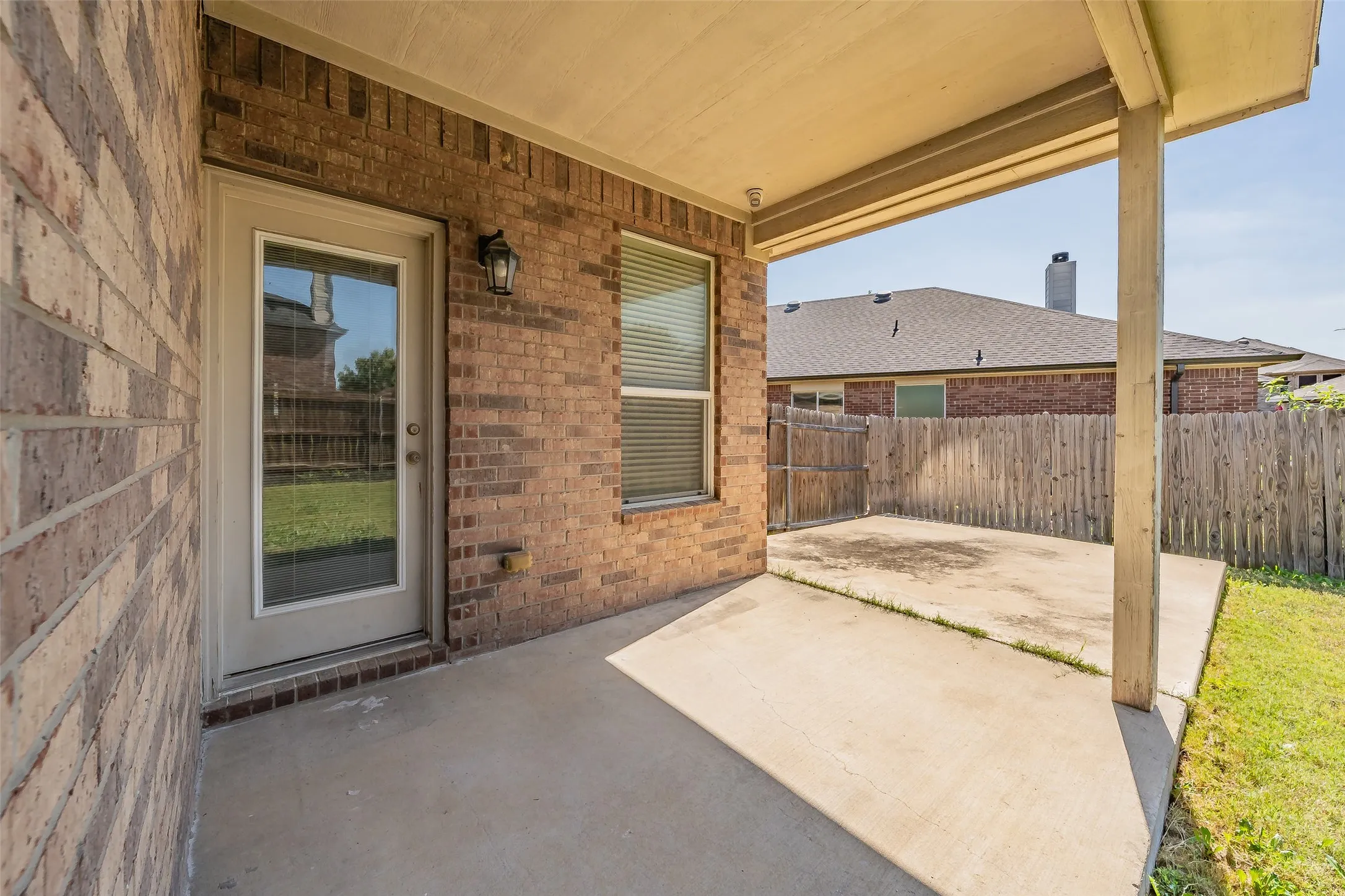 Covered concrete patio with brick exterior, featuring an exterior door with integrated window blinds and an exterior light fixture