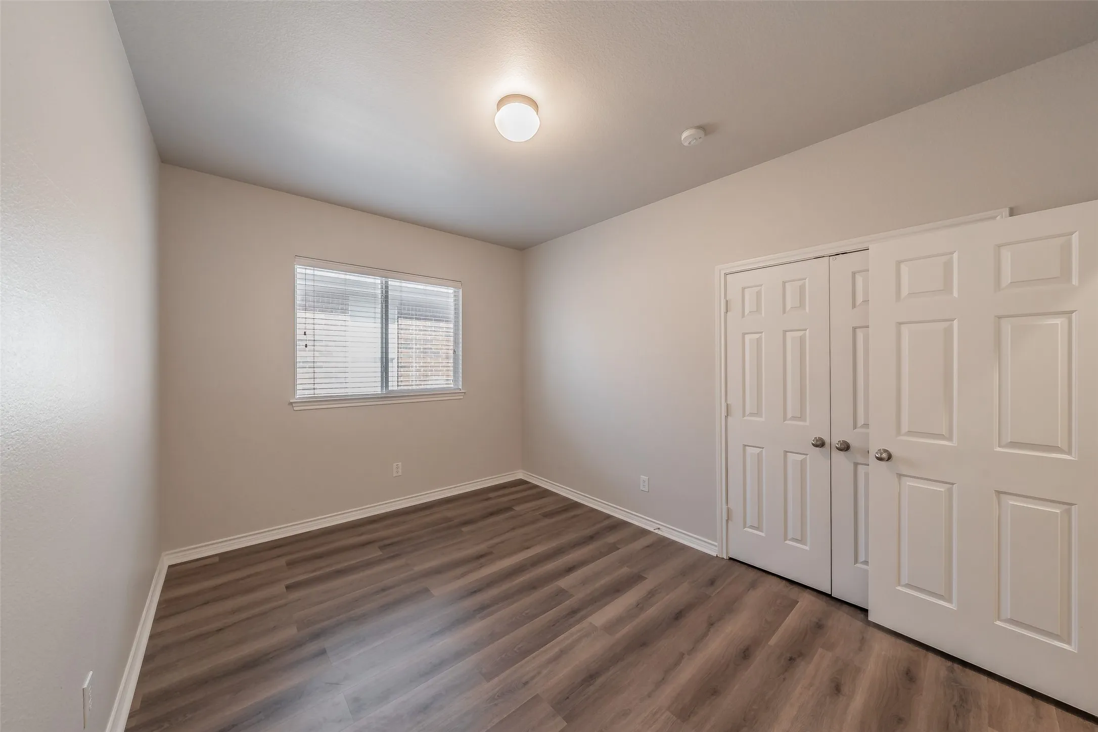 Wood-finish flooring, white trim, window with blinds, and paneled bifold closet doors