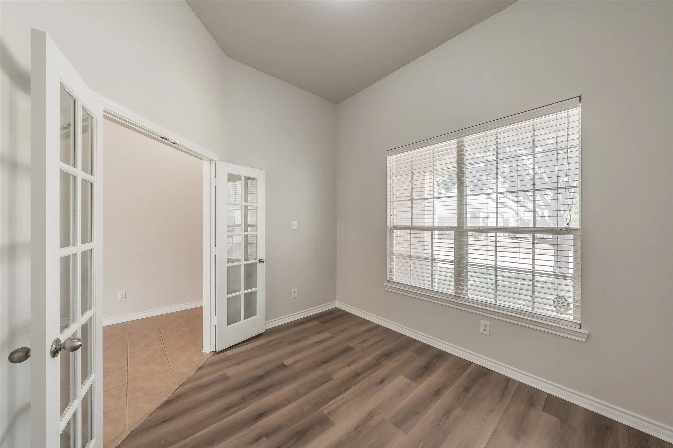 Vaulted ceiling, wood-finish flooring, white trim, window with blinds, and French doors