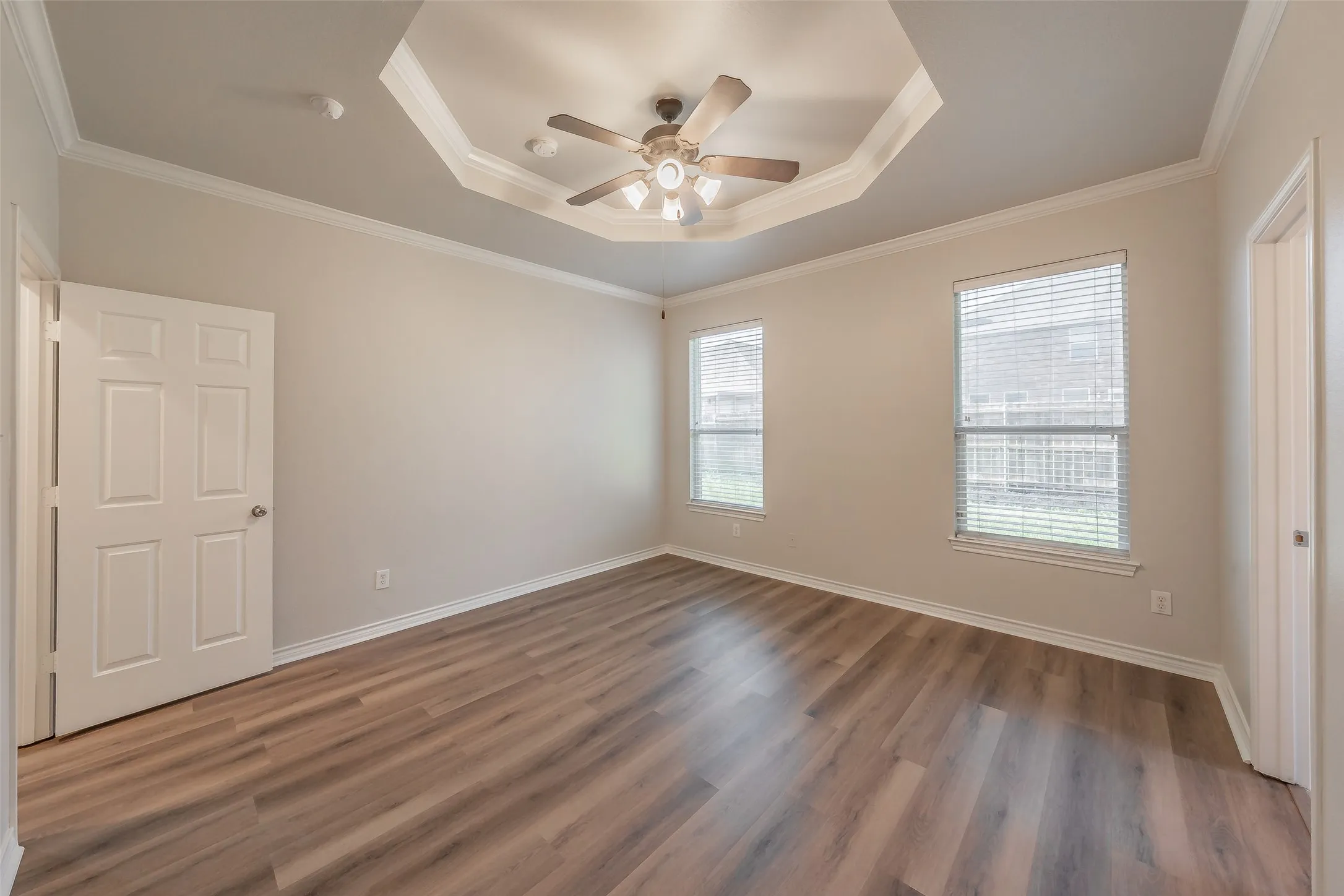Wood-finish flooring, tray ceiling with a ceiling fan, two windows with blinds, and a paneled door