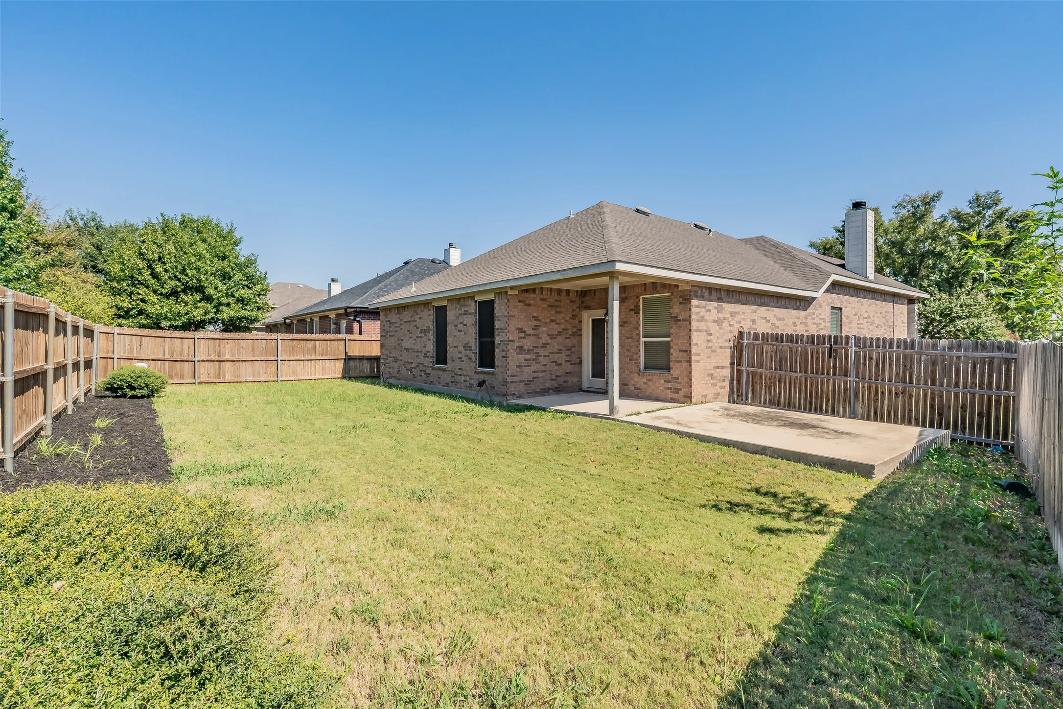 Fenced backyard featuring a covered patio, additional concrete pad, brick exterior, and a shingle roof