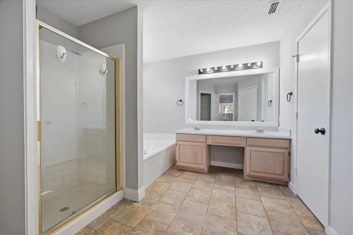 Spa-inspired primary bathroom featuring a spacious walk-in shower with glass enclosure and brushed brass fixtures, dual-sink vanity with wood-finish cabinetry and quartz countertop, illuminated mirror with integrated lighting, and neutral tile flooring throughout